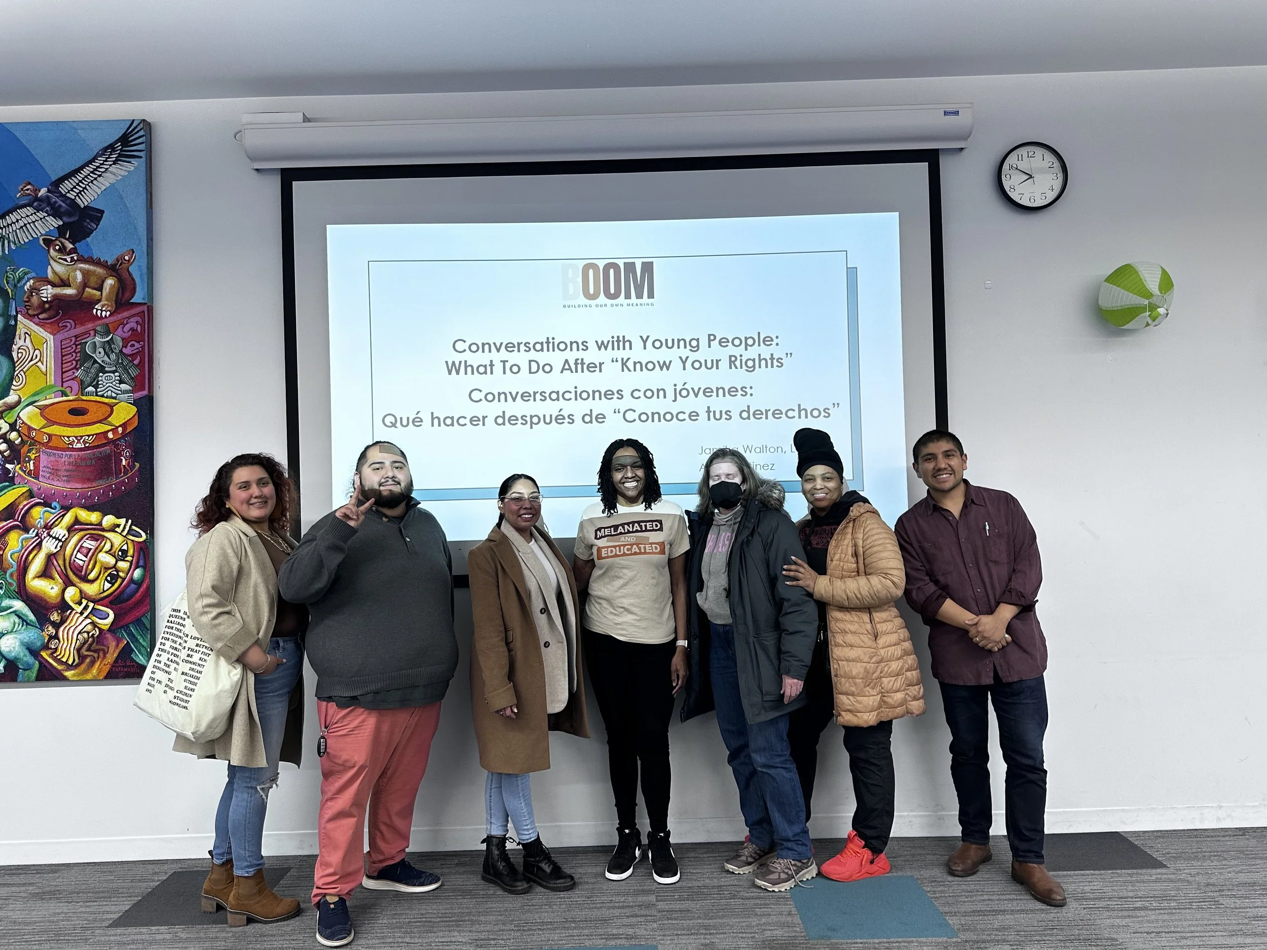 A group of eight diverse people standing in front of a presentation screen at an indoor event. The screen displays a presentation titled 'Conversations with Young People: What To Do After Know Your Rights'. The group is smiling, some with arms around