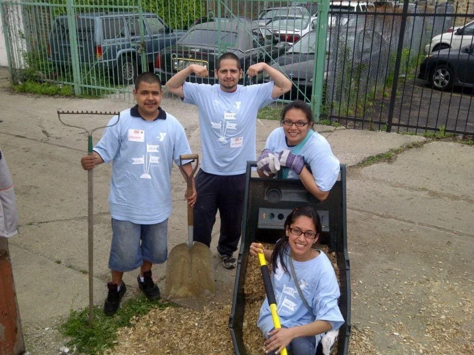 Group of four volunteers at a community cleanup event. One person holds a rake, another a shovel, and a third is sitting in a trash bin holding a broom. The group is smiling and wearing light blue T-shirts with a logo. There is a fence and parked car