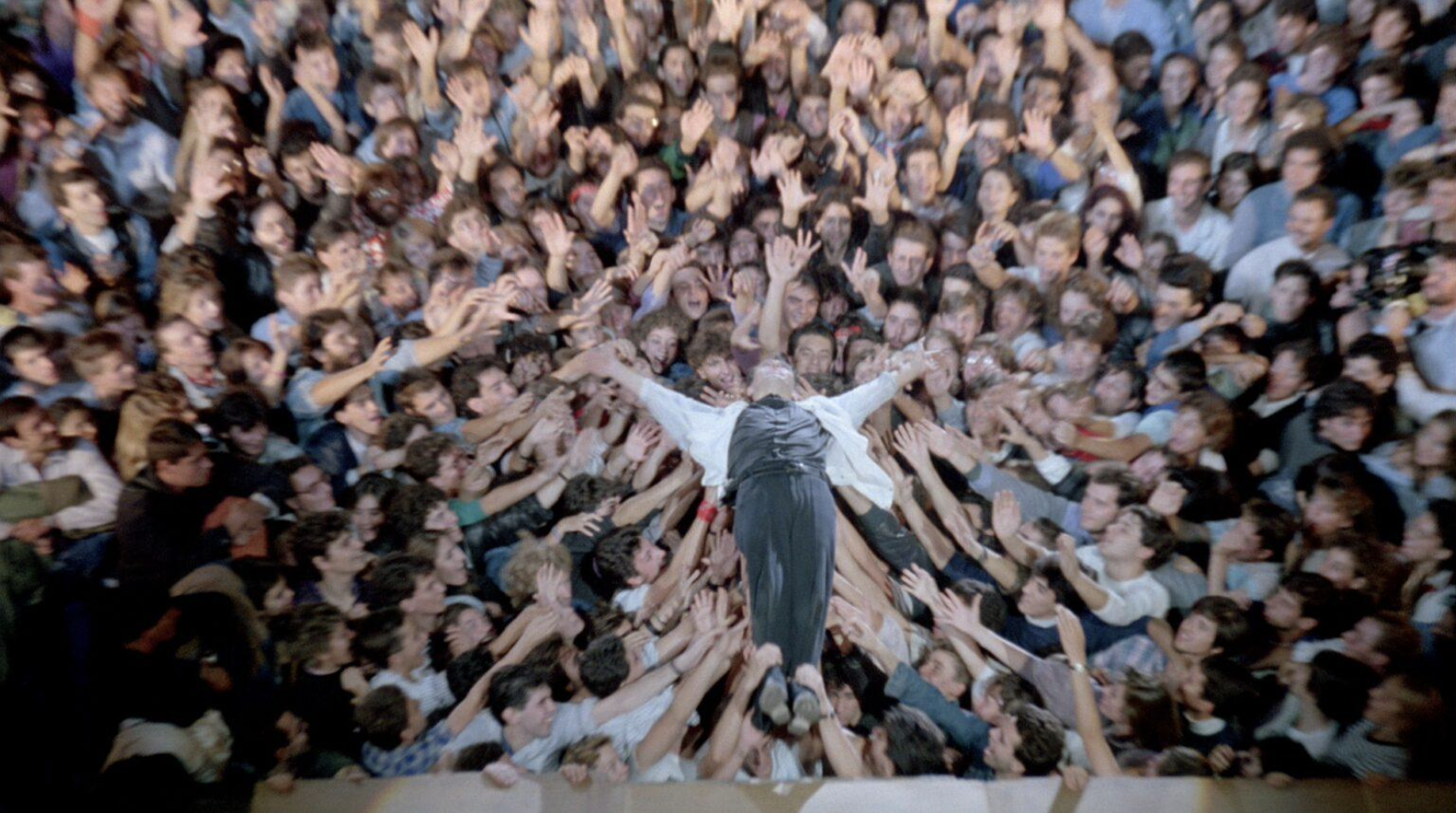 A person in a white shirt and black pants is stretched out with arms wide open, lying on a surface being reached out to by many people surrounding them, creating a crowd scene with excitement and enthusiasm.