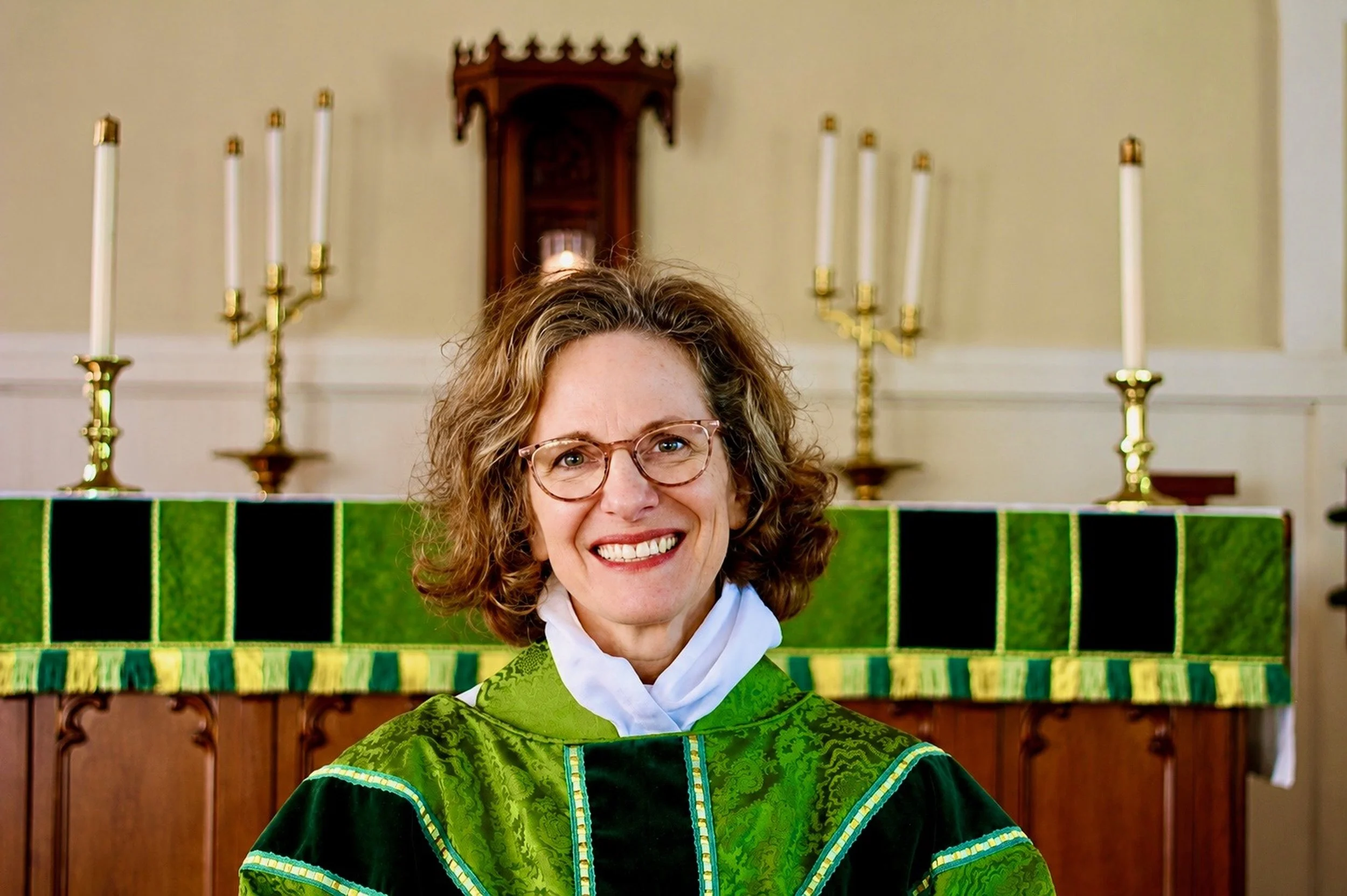 A woman with curly hair and glasses smiling, wearing a green academic gown, standing in front of a church altar with candelabras and candles visible in the background.