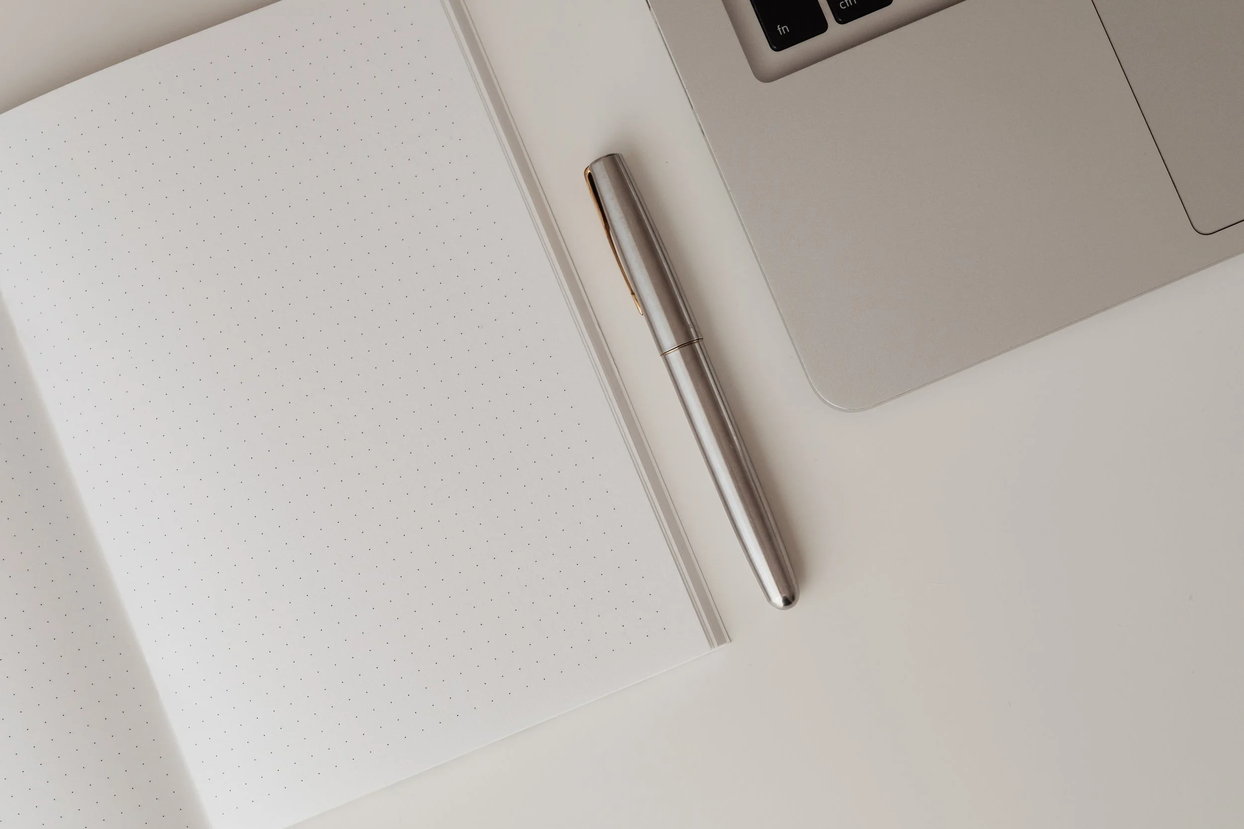 Silver pen on an open dotted notebook beside a laptop.