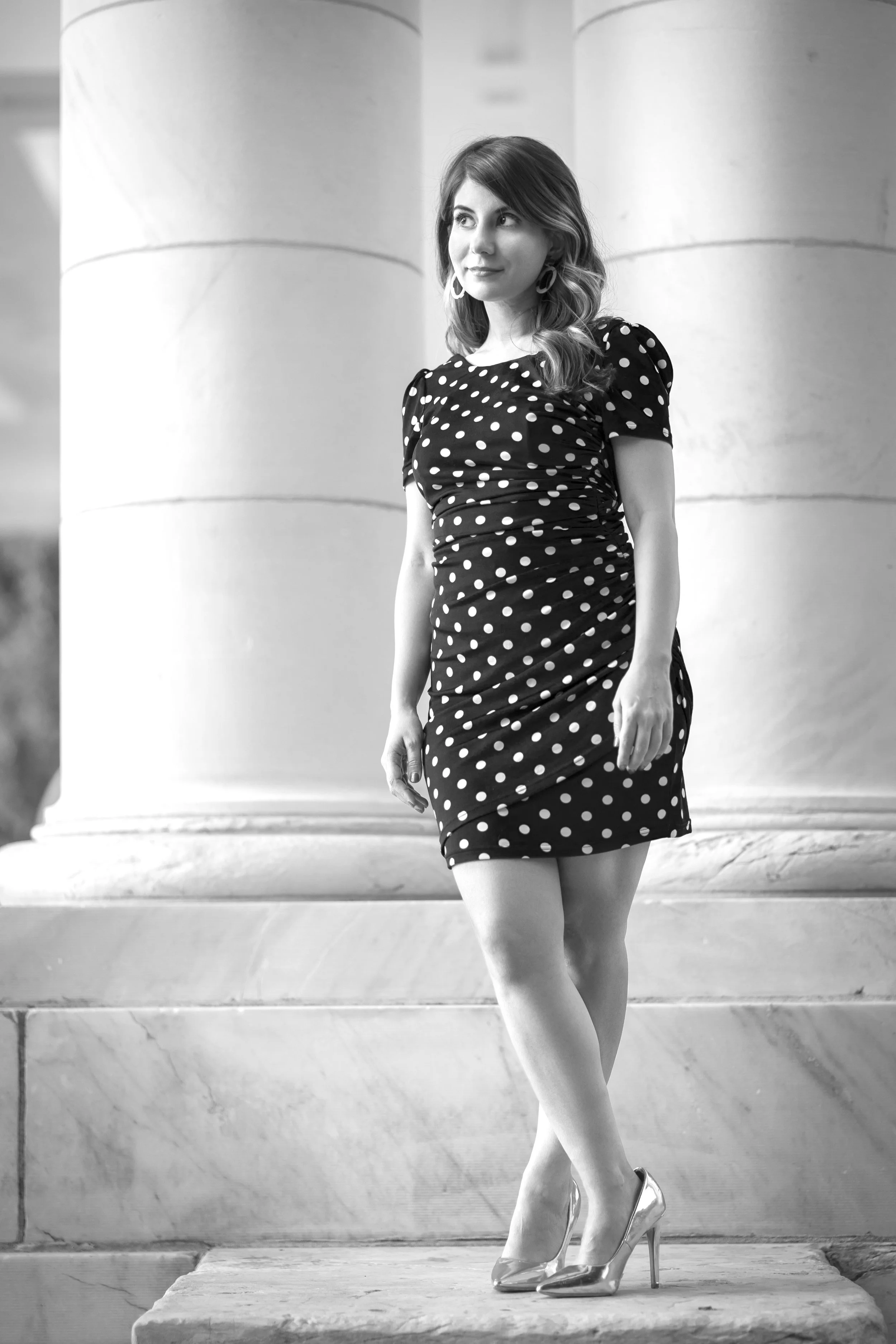 A beautiful woman in a polka dot dress and high heels standing on stairs, with large column-like pillars in the background at Cheesman Park in Denver, CO during a creative and professional photoshoot for new portraits.
