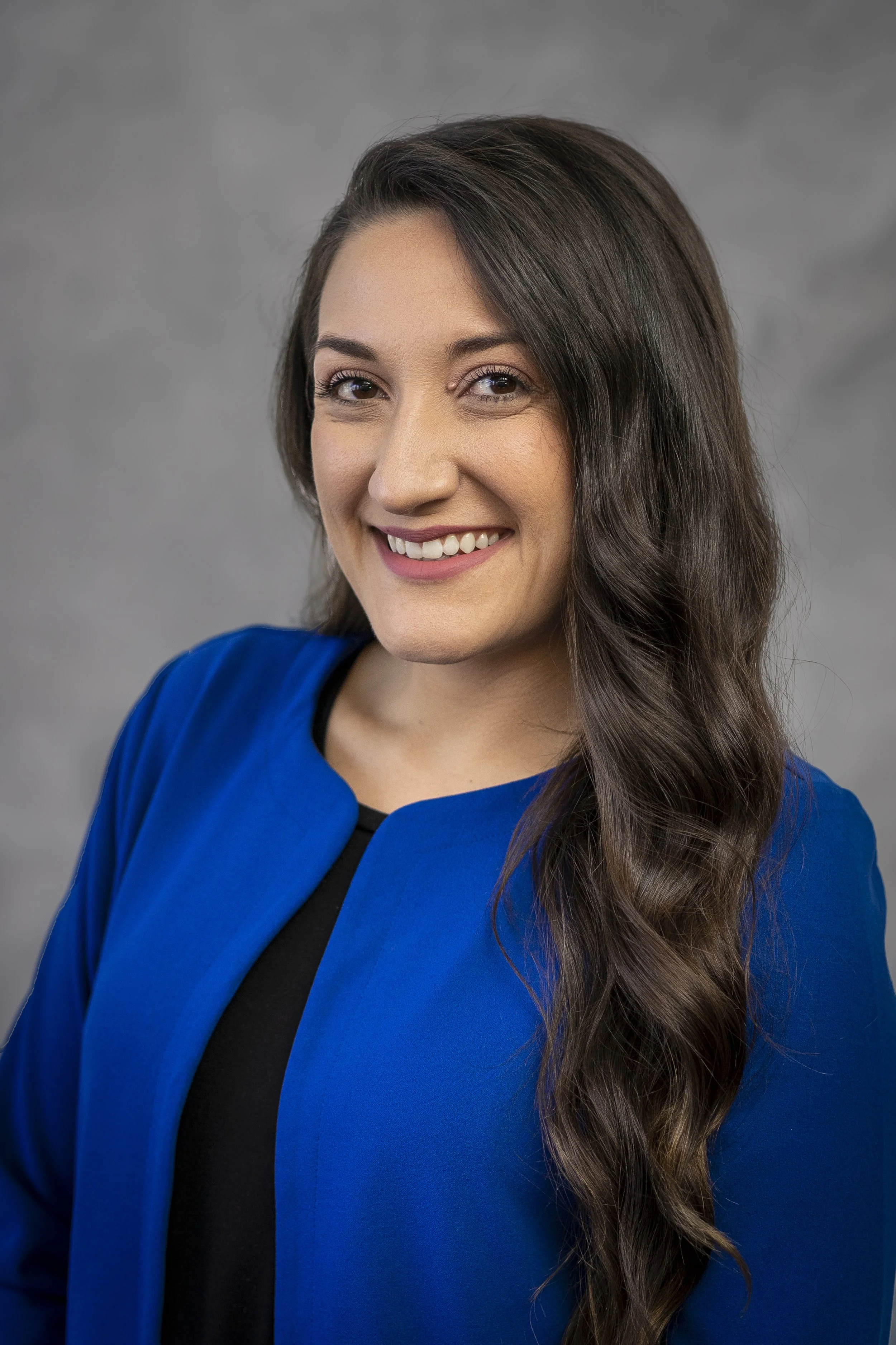 A professional and corporate headshot of a woman with long wavy dark brown hair, wearing a blue blazer over a black top, smiling at the camera against a gray background.