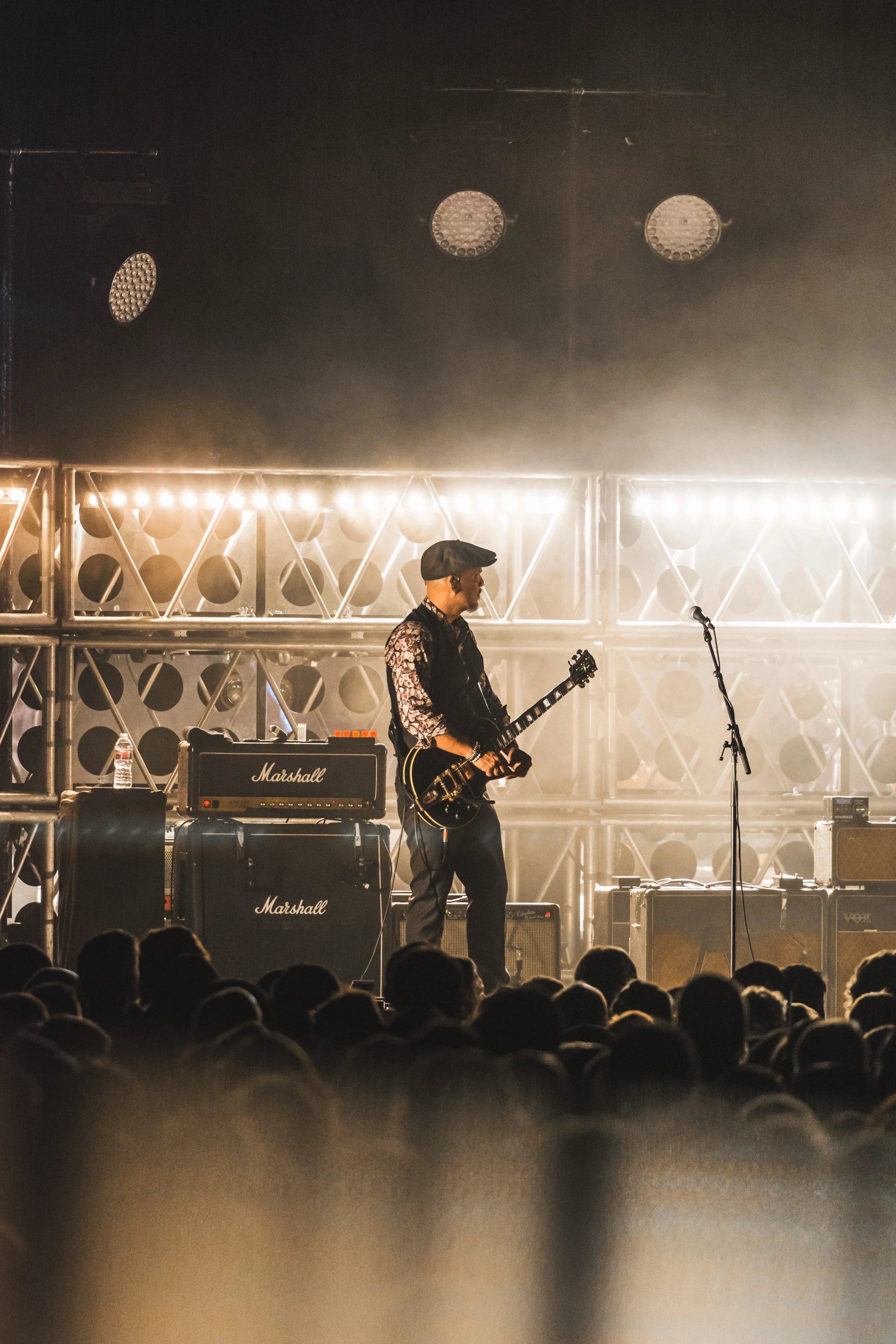 A musician with the band The Pixies wearing a flat cap and floral shirt playing an electric guitar on stage in front of an audience. The stage features Marshall amplifiers and metal scaffolding illuminated by bright stage lights.