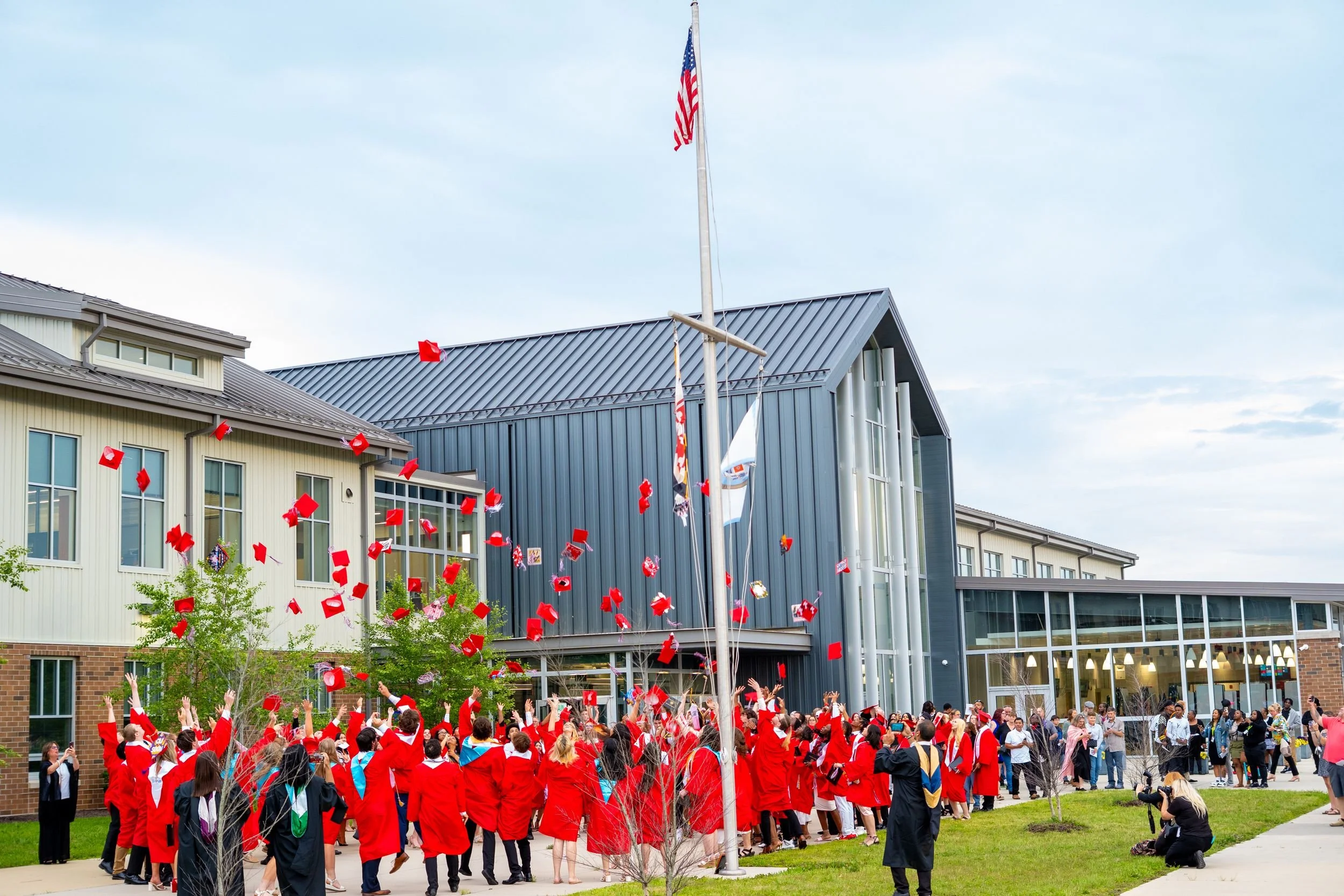 Graduating students in red caps and gowns toss their caps outside a modern school building during a graduation celebration.