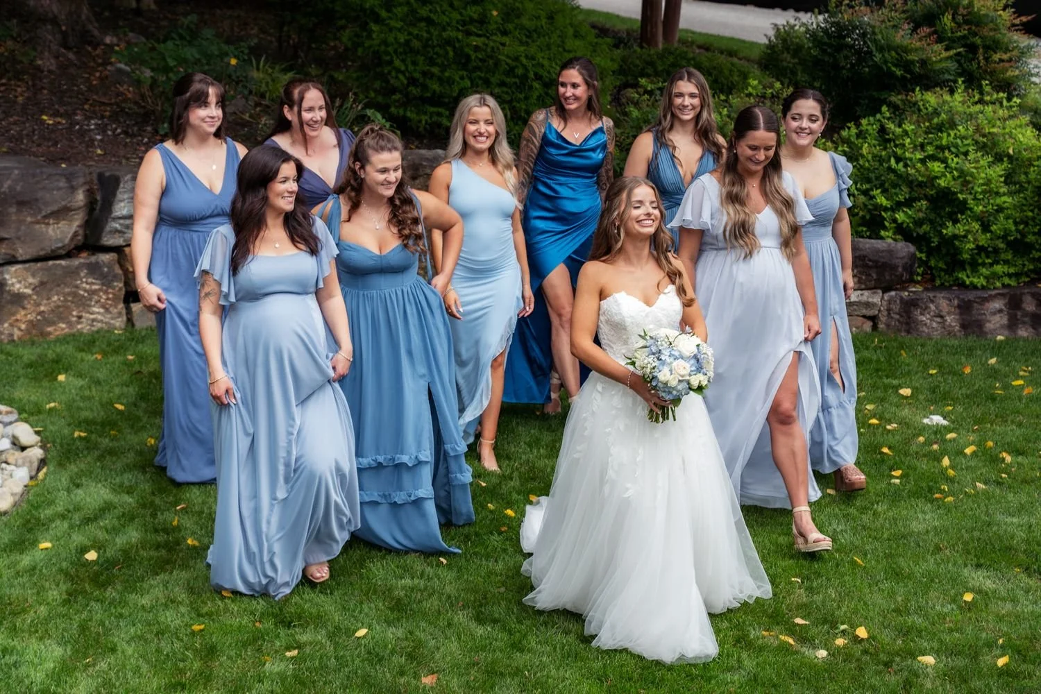 Bride and bridesmaids in blue dresses smile during outdoor wedding party portraits with a white and blue bouquet.