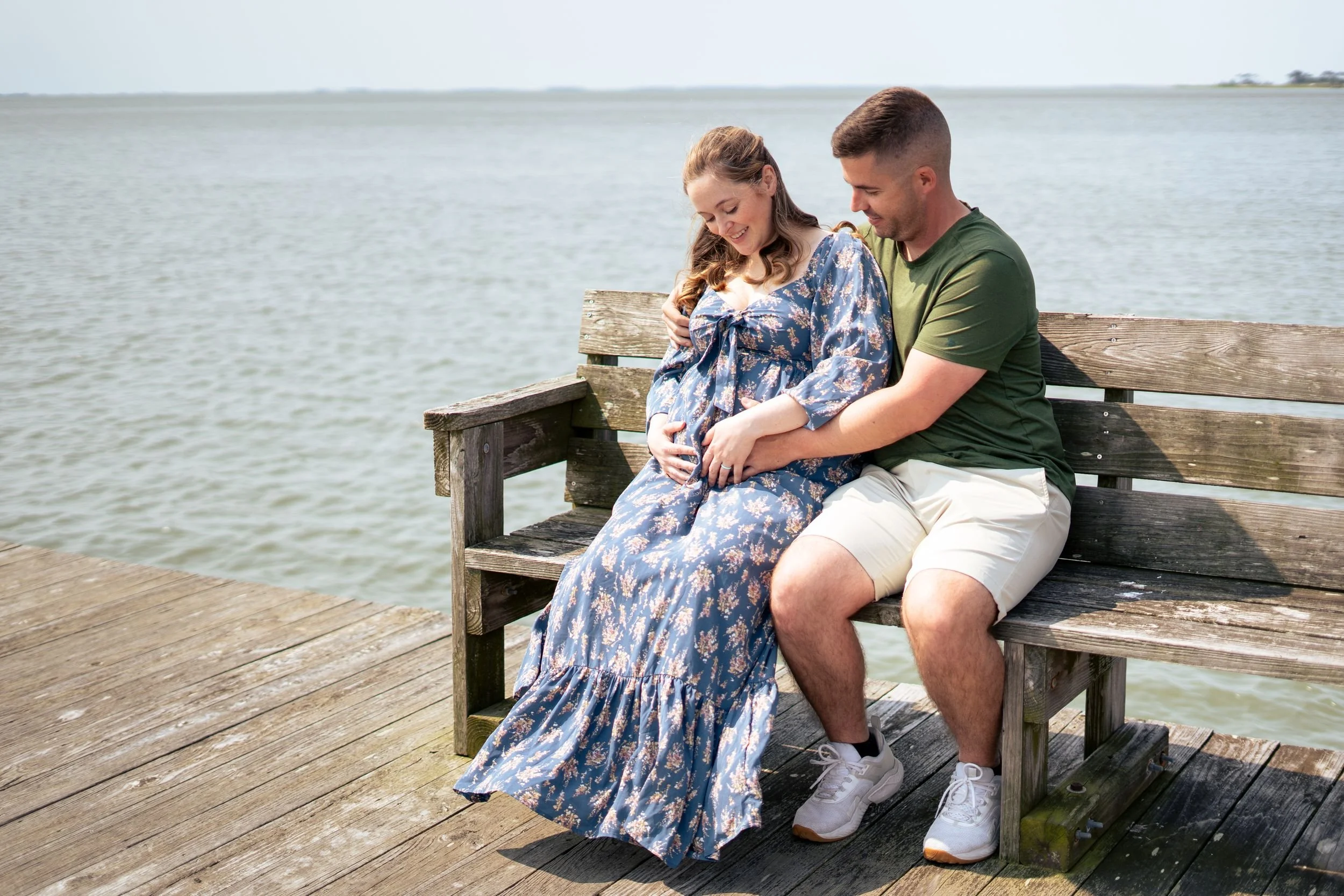 Expecting parents pose for maternity photos on a wooden bench by the lake.