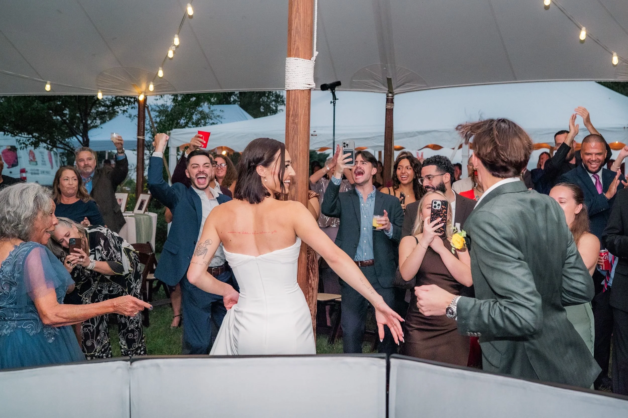 Bride and groom celebrate their grand entrance with cheering guests at an outdoor wedding reception under a canopy with string lights.