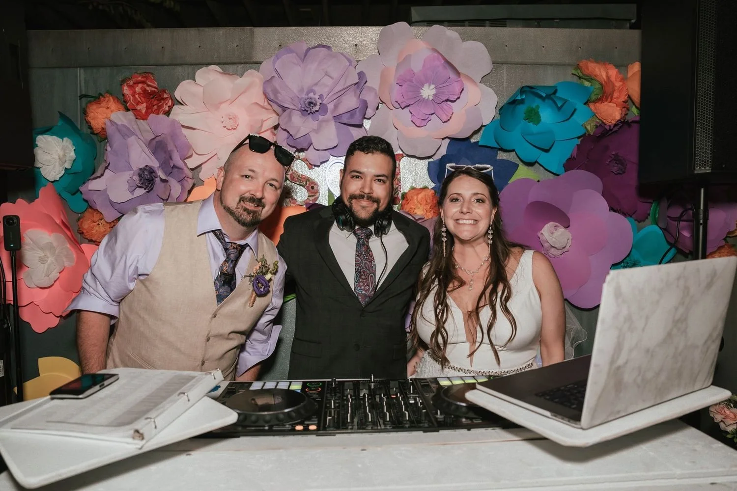 dj and bride and groom smiling at a celebration, standing behind a DJ setup with a laptop and mixer, in front of a large colorful paper flower backdrop.