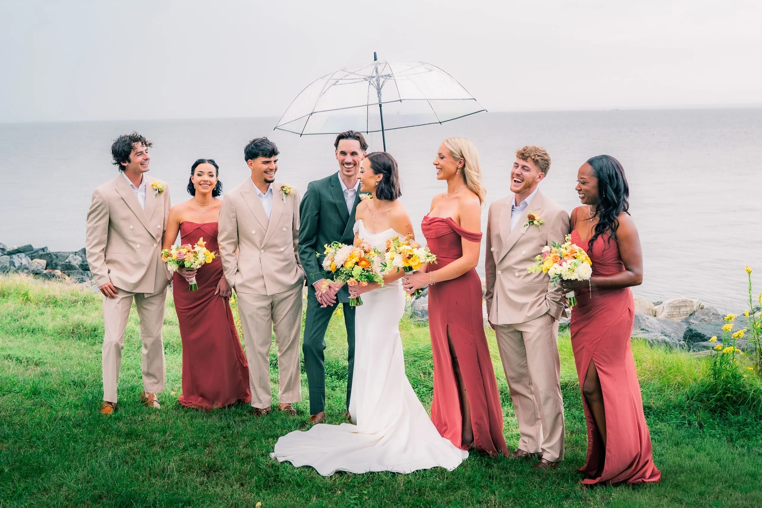 A wedding party standing outdoors by the water, with a bride and groom in the center, holding bouquets, and surrounded by bridesmaids and groomsmen, some of whom are holding an umbrella.
