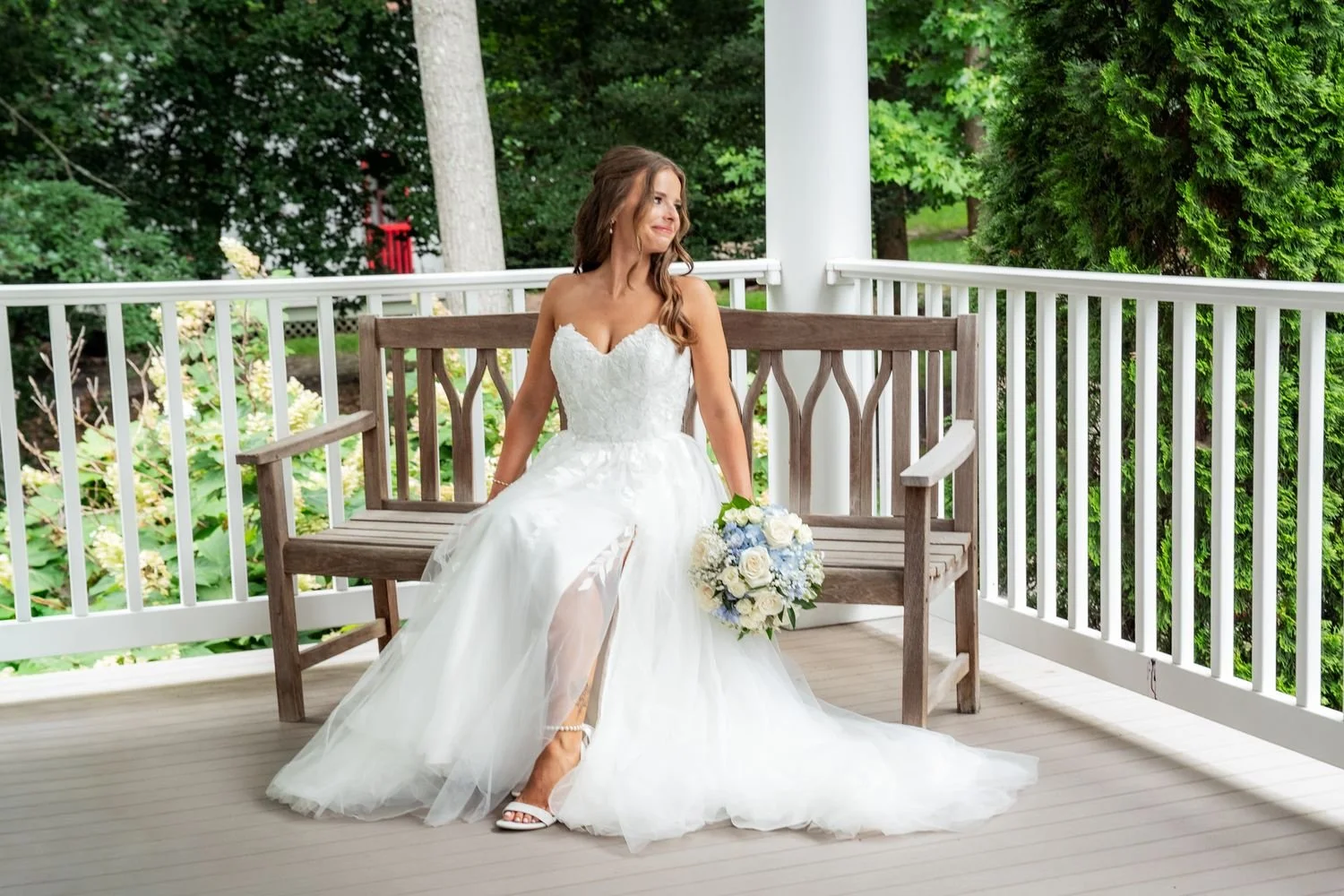 Bride in a white wedding gown sitting on a wooden bench on a porch, holding a bouquet of white and blue flowers, with greenery in the background.