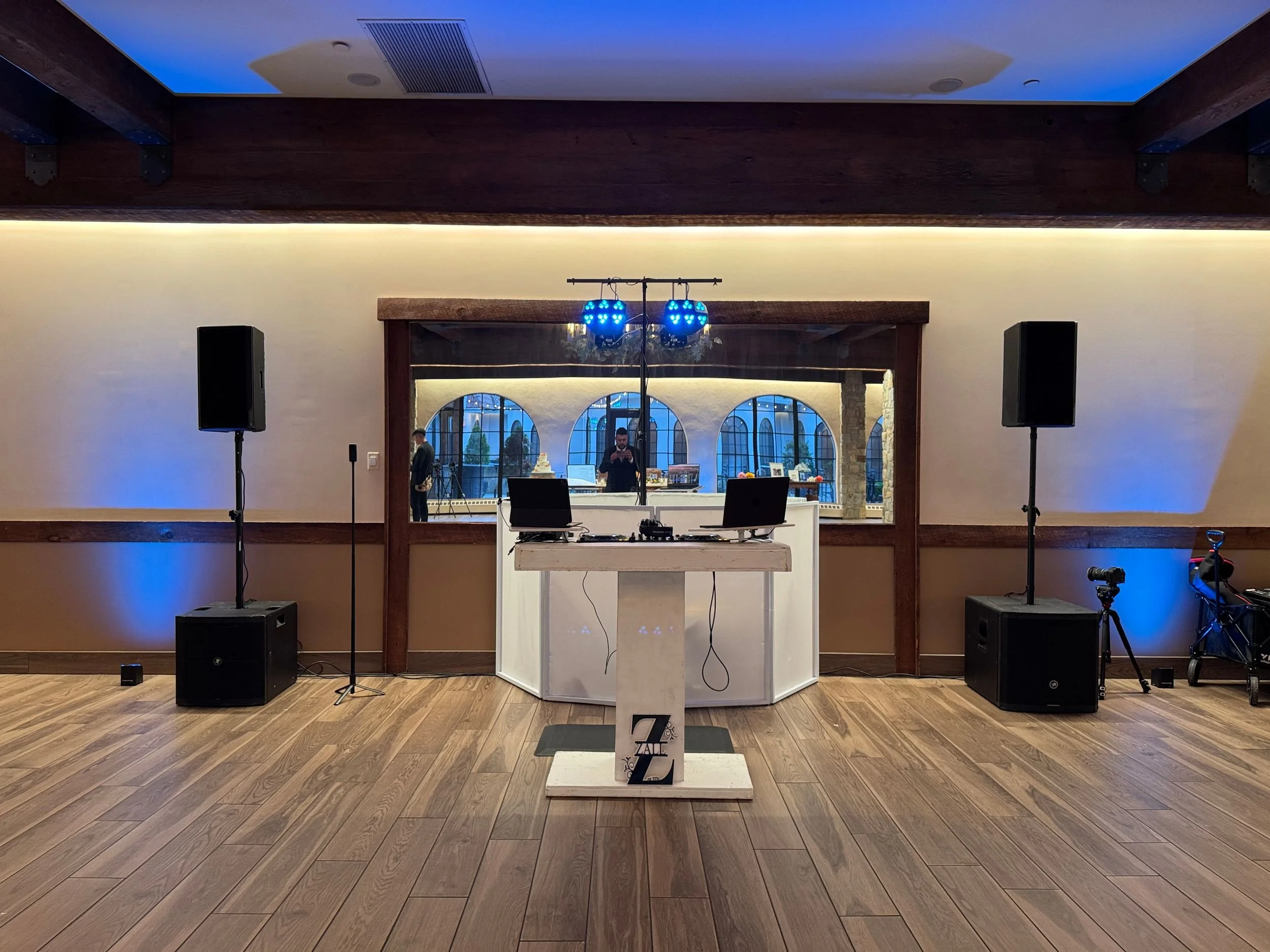 Indoor event space with a DJ booth, blue disco lights, and speakers. Behind the booth, people are visible through a window with arched frames. The room has wooden flooring and a ceiling with blue lighting.