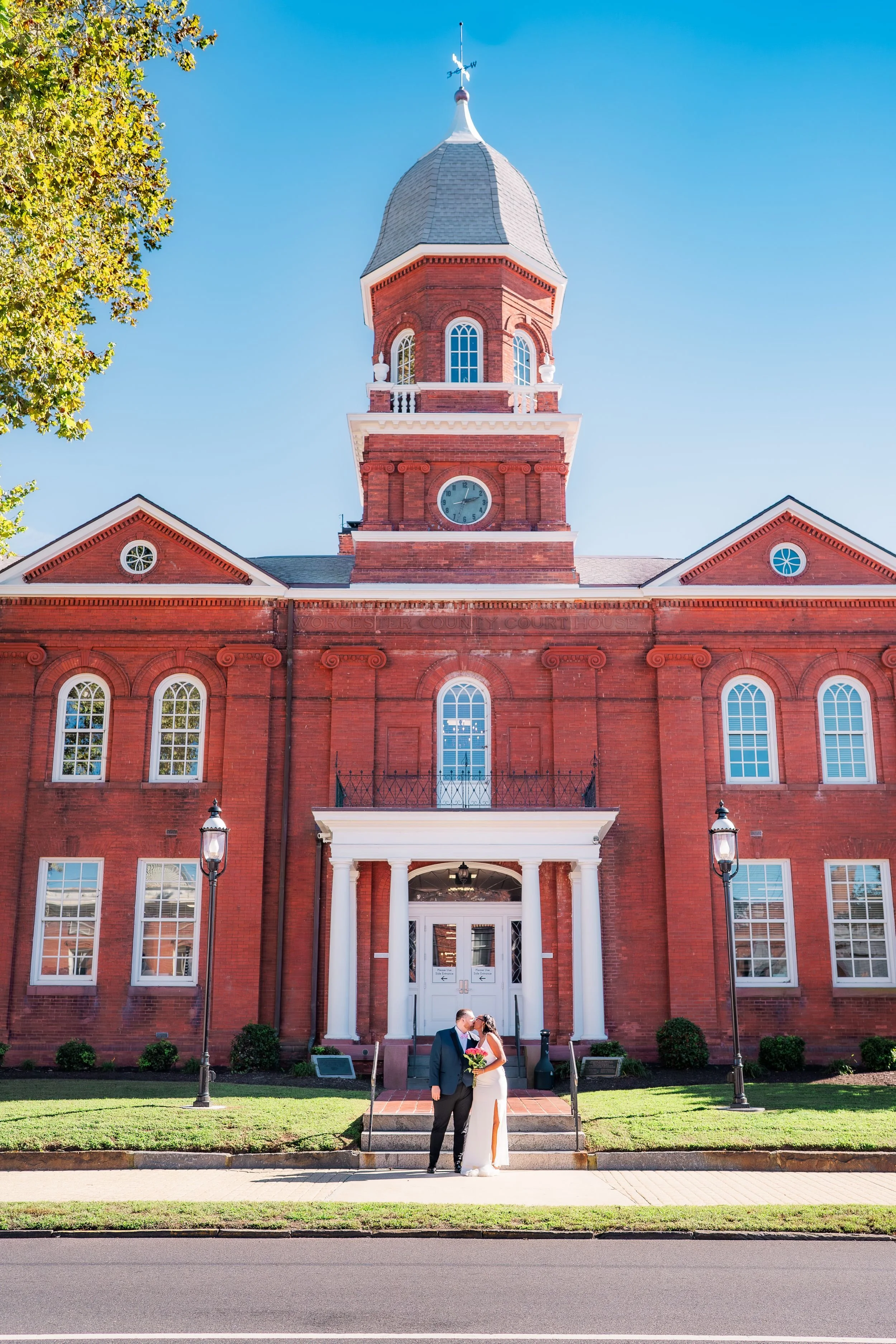 A bride and groom kissing in front of a brick courthouse on a sunny day. The bride is holding a bouquet, and the groom is dressed in a dark suit. The courthouse has a clock, arched windows, and a prominent tower with a weather vane on top.