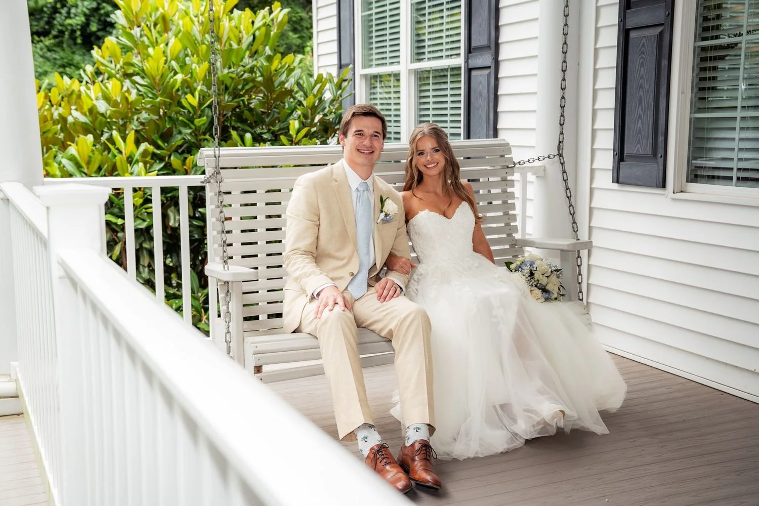 A smiling bride and groom sitting on a white porch swing, with lush greenery in the background. The bride is wearing a white wedding dress and holding a bouquet, while the groom is dressed in a beige suit with a light blue tie.