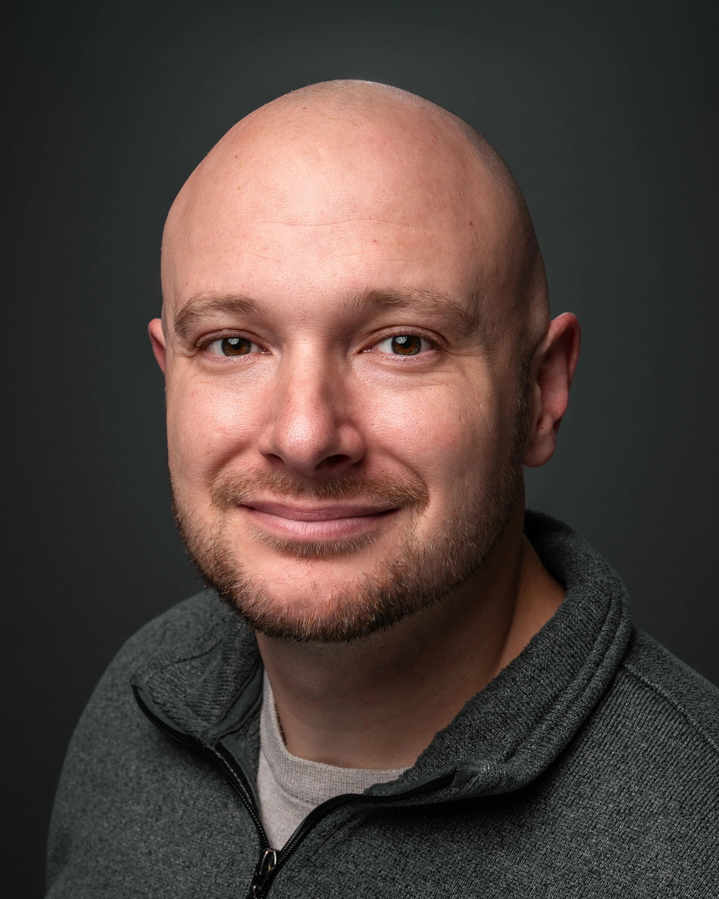 Close-up of a smiling man with a shaved head, light beard, and brown eyes, wearing a gray hoodie on a dark background.