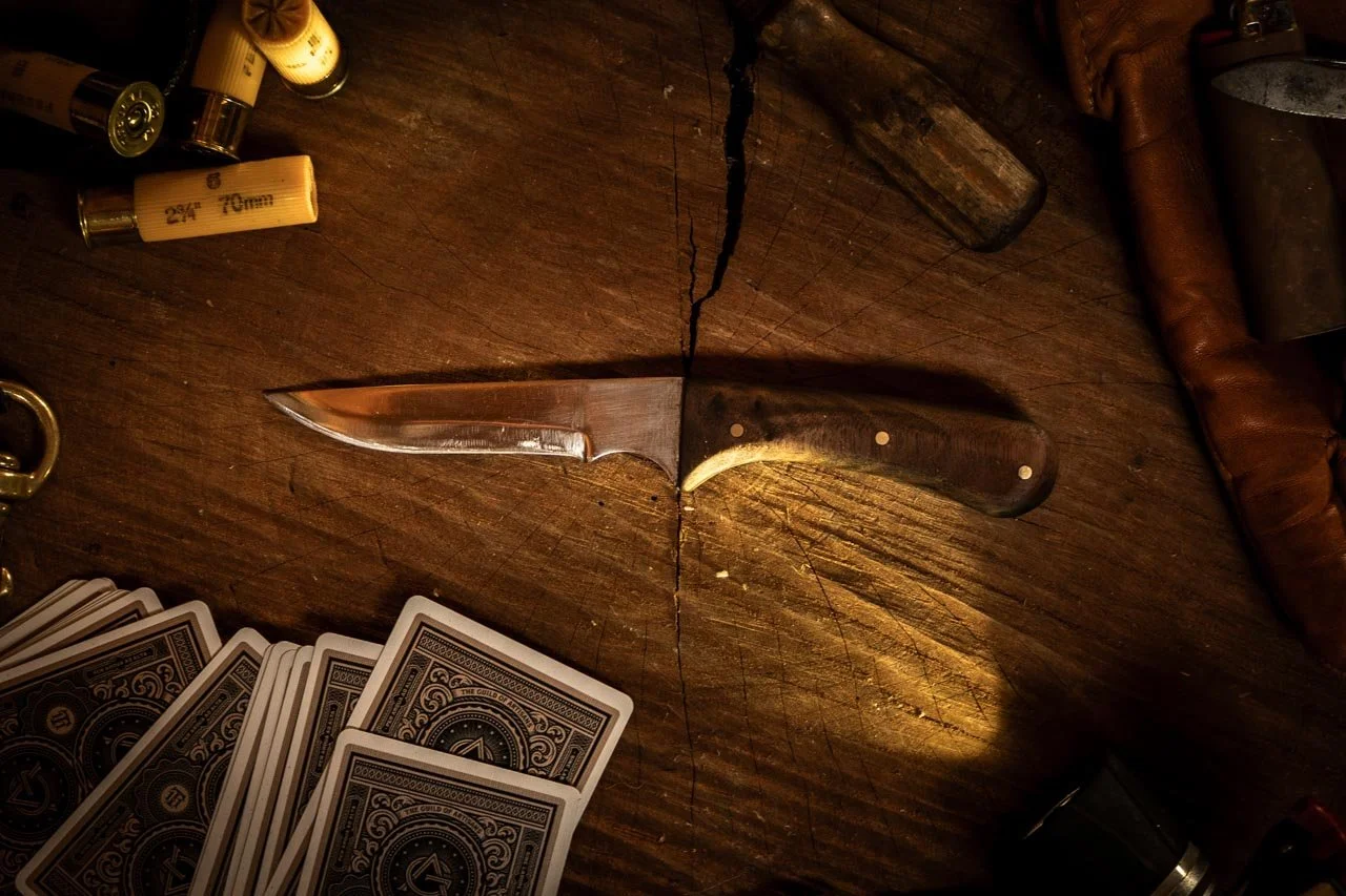 A wooden table with a hunting knife in the center, playing cards, bullets, and metal objects surrounding it.