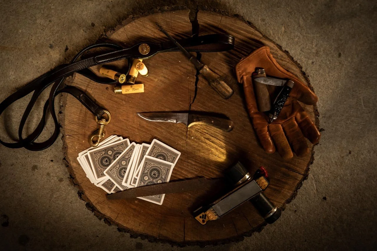 Items placed on a tree stump include a deck of playing cards, a leather glove with a folding knife, a pistol with a flashlight, a small black-and-yellow tool, a compact flashlight, a lanyard with bullets, and a twisting knife.