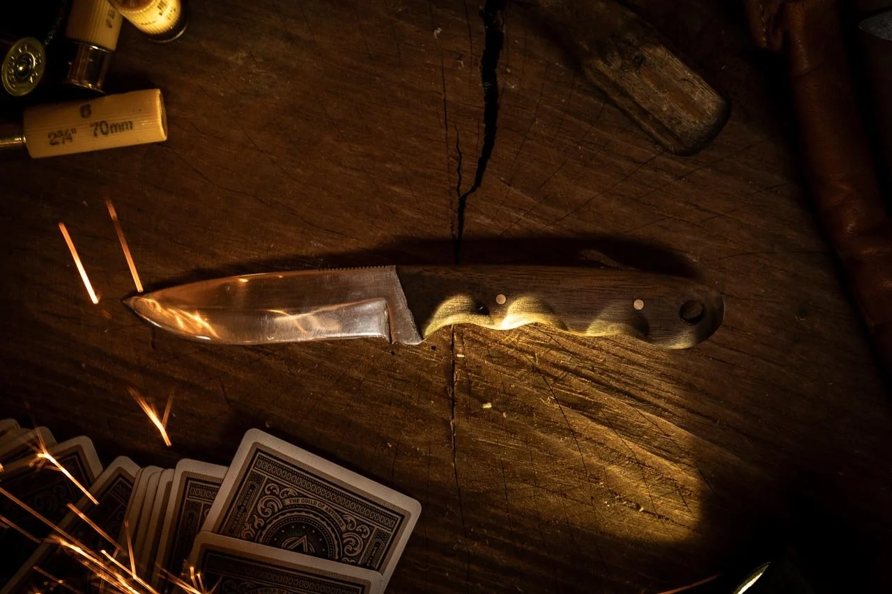 A rustic wooden table with a hunting knife, playing cards in borders, and a few shotgun shells surrounding it.