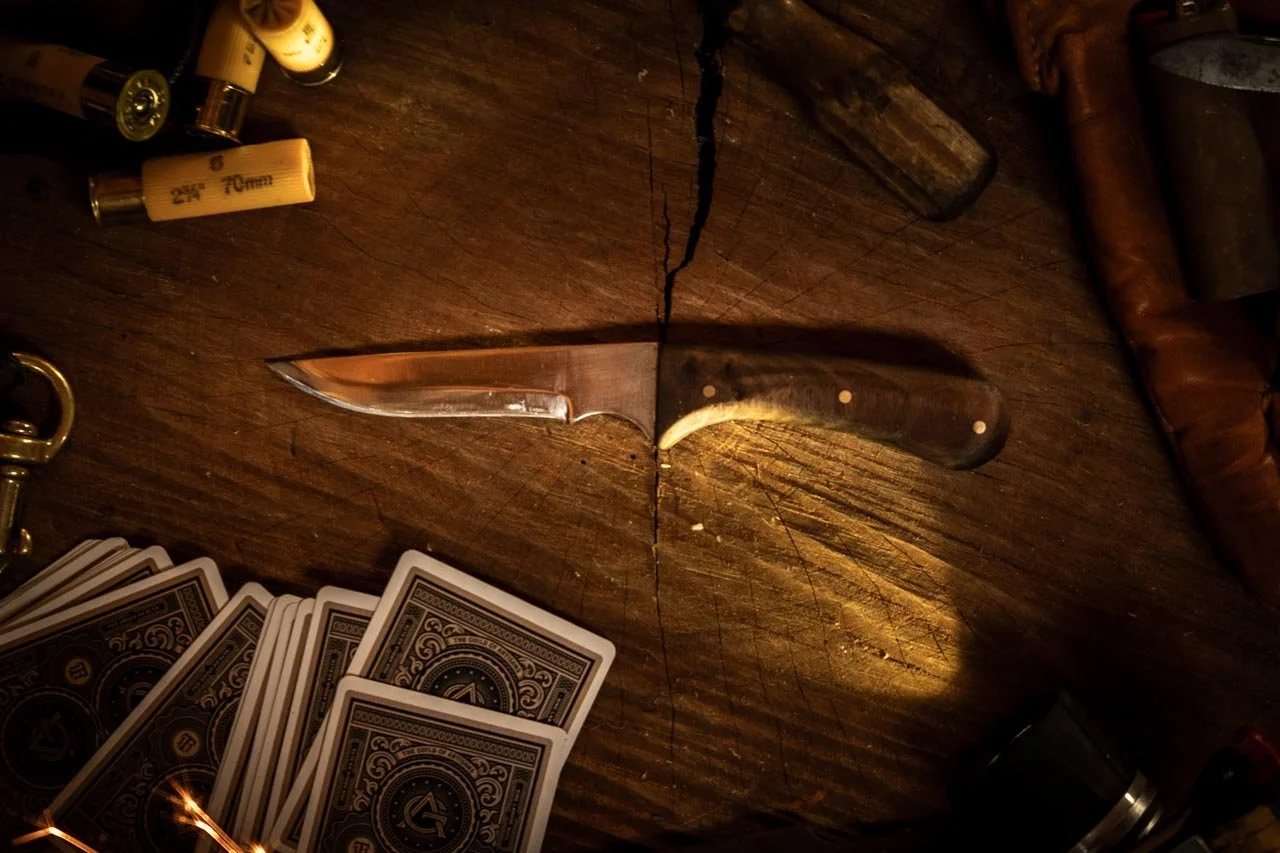 A knife with a wooden handle and a sharp blade placed on a wooden surface, with playing cards, shotgun shells, and shotguns surrounds it.