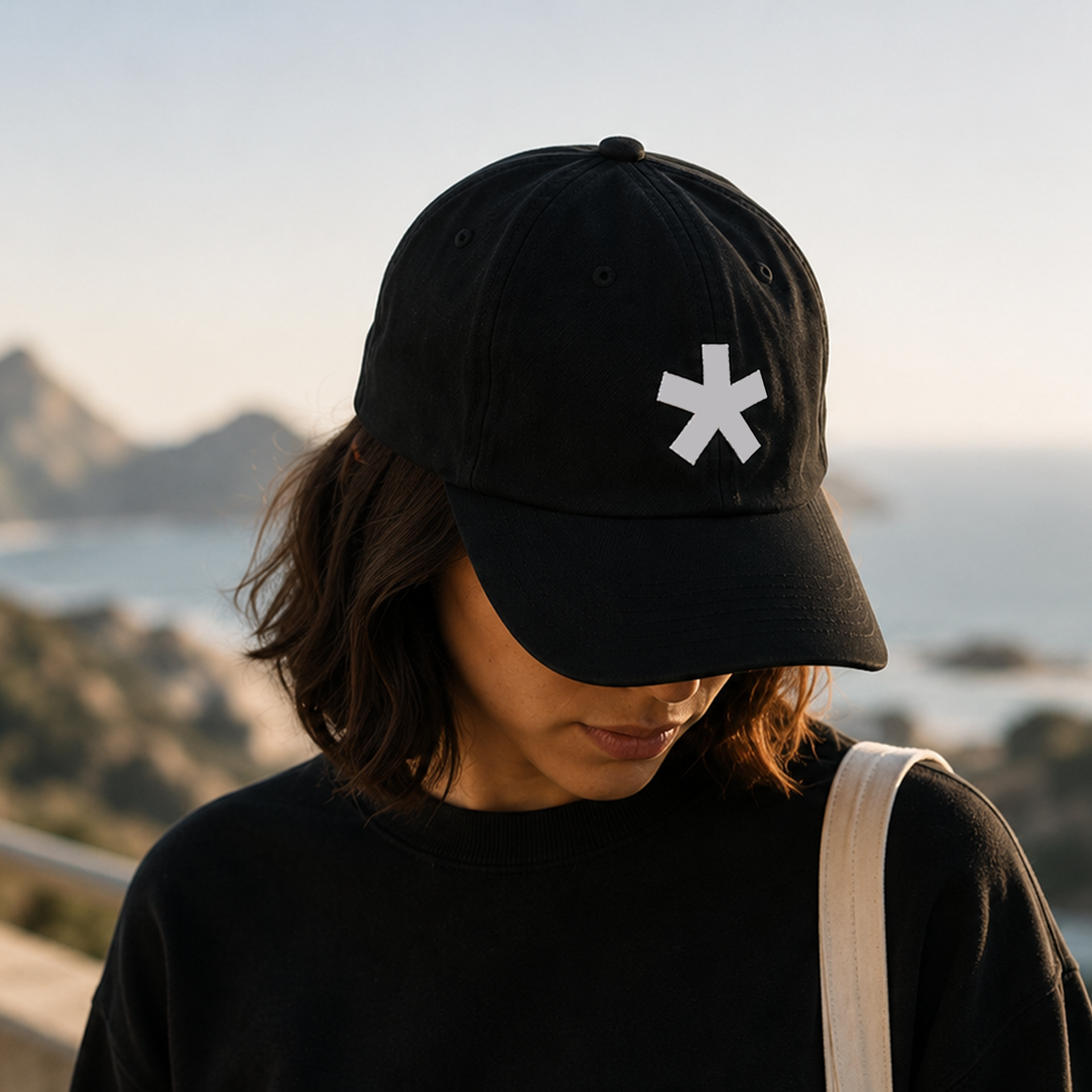 Person wearing a black baseball cap with a white asterisk logo, with brown hair, outdoors at sunset, with mountains in the background.