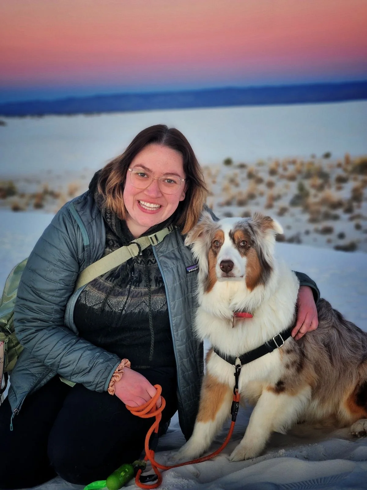 dog parent Jesse and their dog in front of a sunset and white sand. Jesse has medium-length brown hair, wears glasses with transparent frames. Basil is a white and tan Australian Shepherd who looks fluffy.