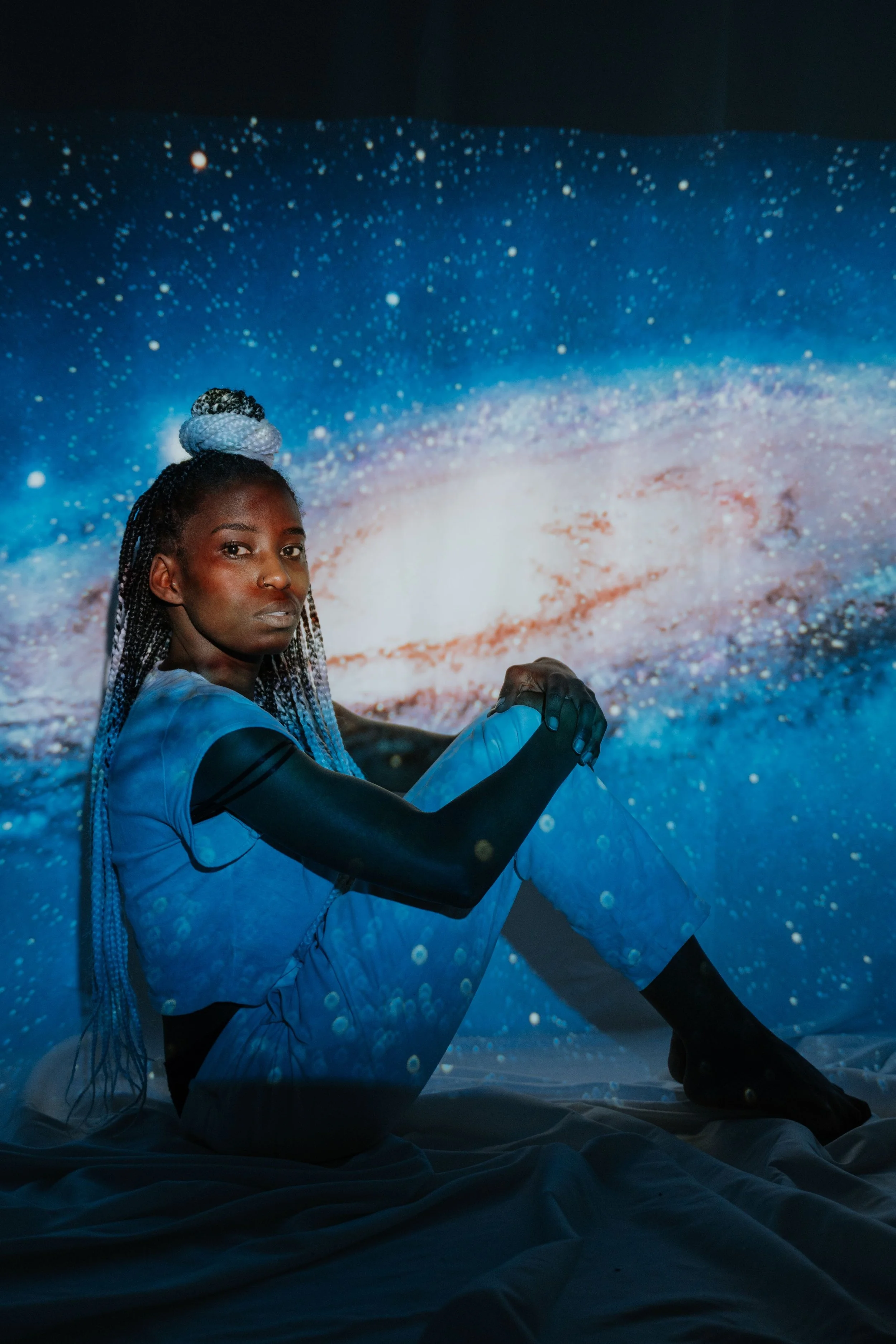 teen with dark skin and long dreads sits with their hands on their knees in a space background