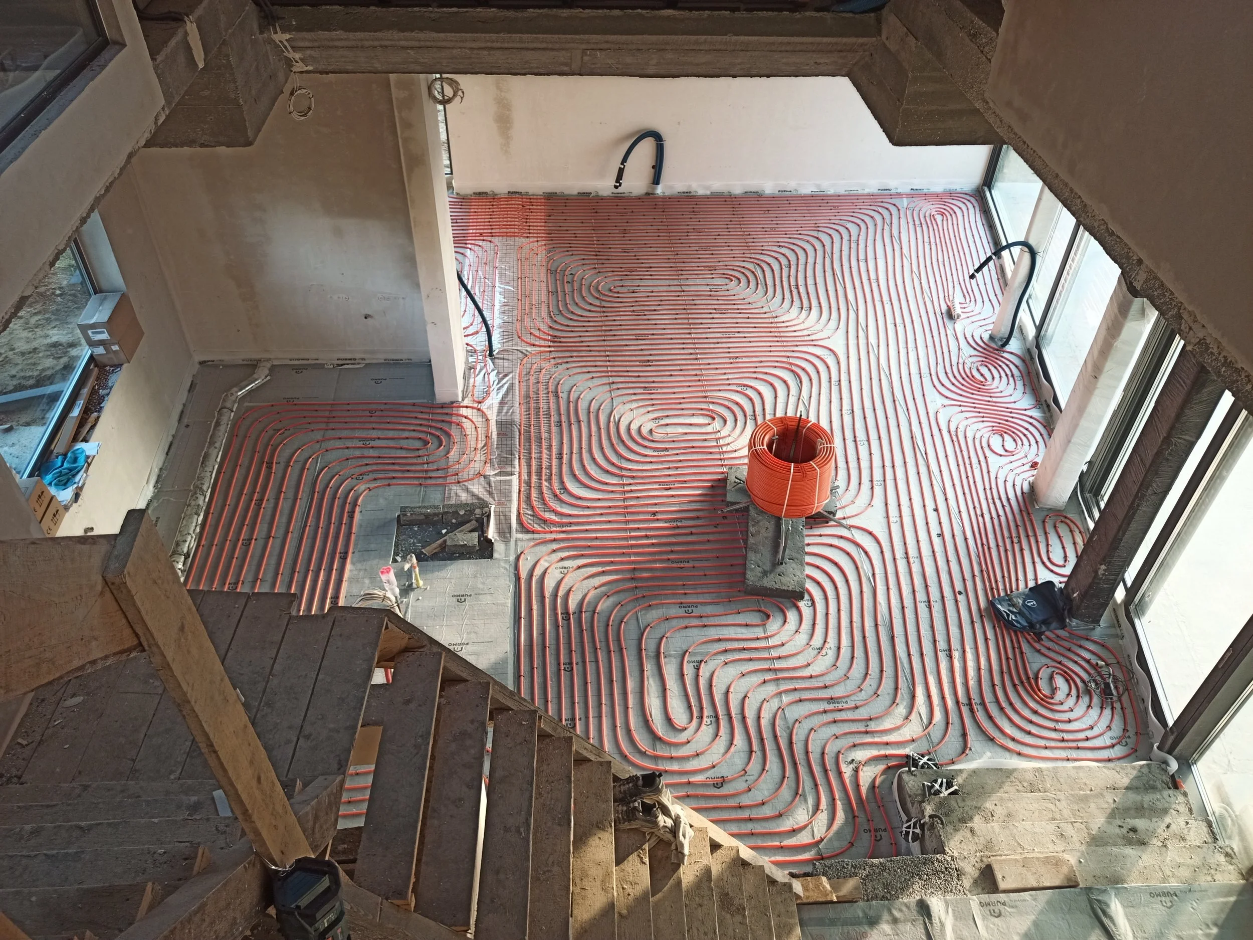 Under construction room with radiant floor heating pipes installed on the floor, visible through large windows, with unfinished walls and stairs in the foreground.