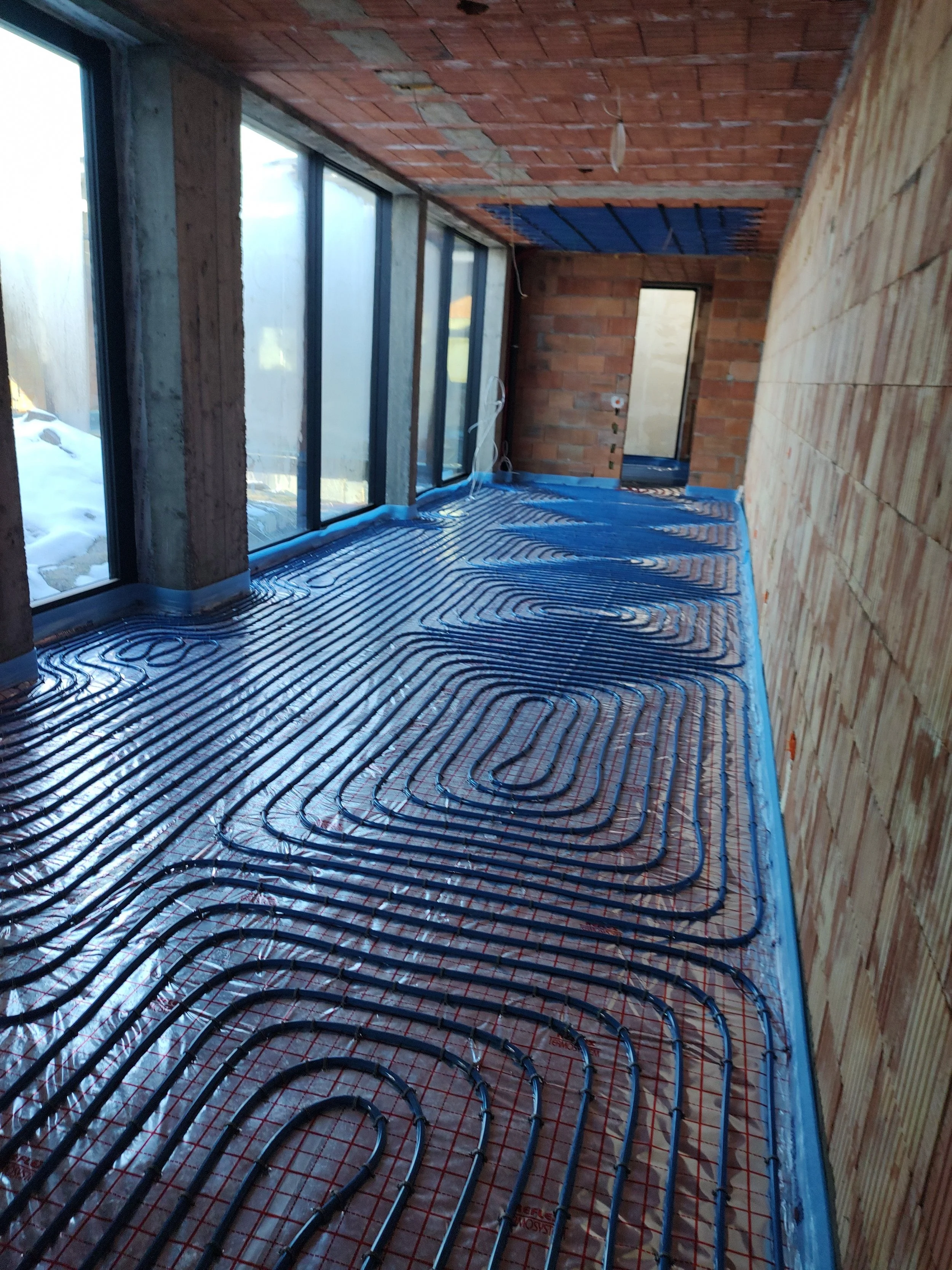 Under construction room with underfloor radiant heating pipes installed, brick walls and large glass windows.