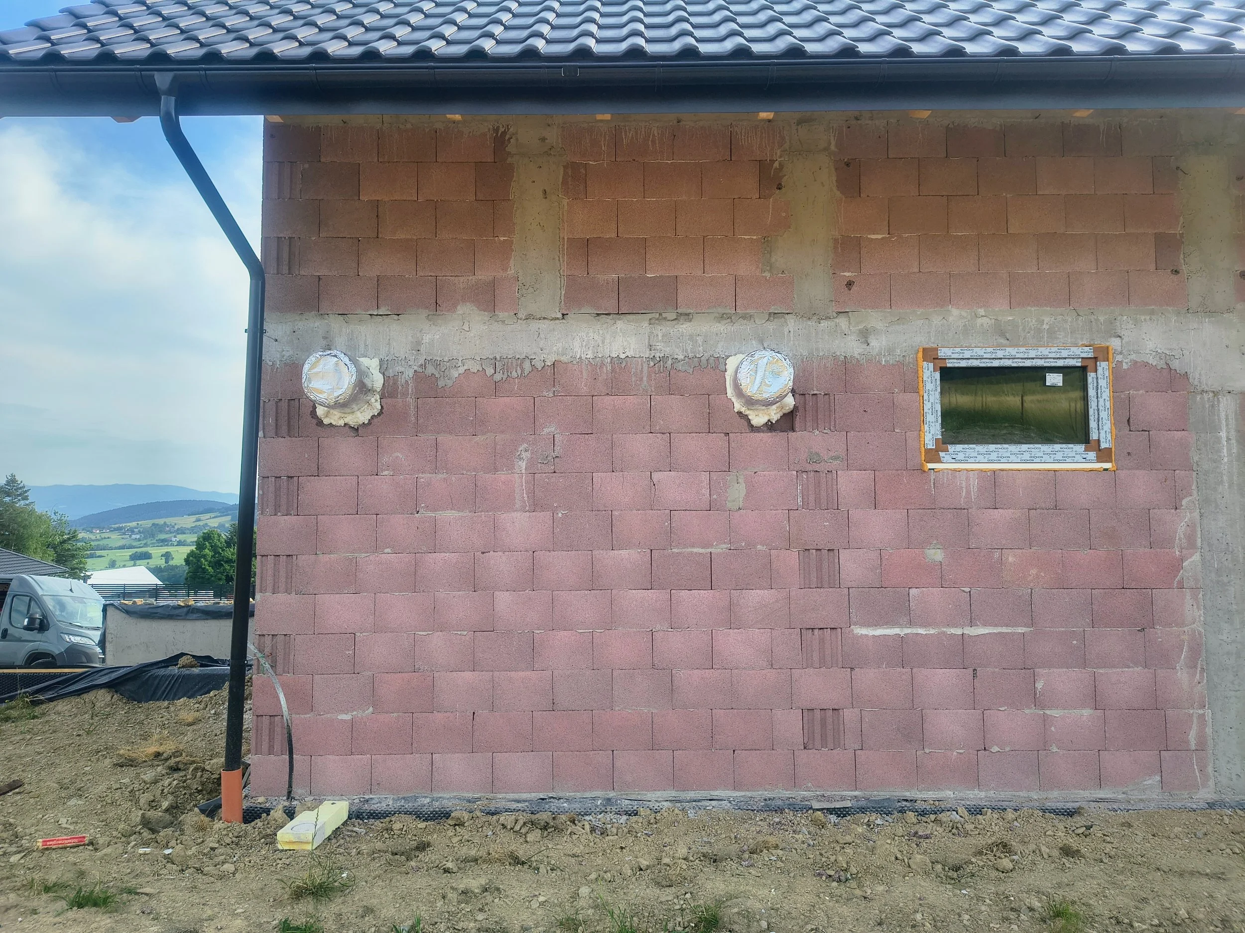 A building under construction with red brick walls, a small rectangular window, and ventilation pipes on the wall. The roof has dark shingles, and there is a black rain gutter along the edge. The ground is bare soil with some grass patches, and a car