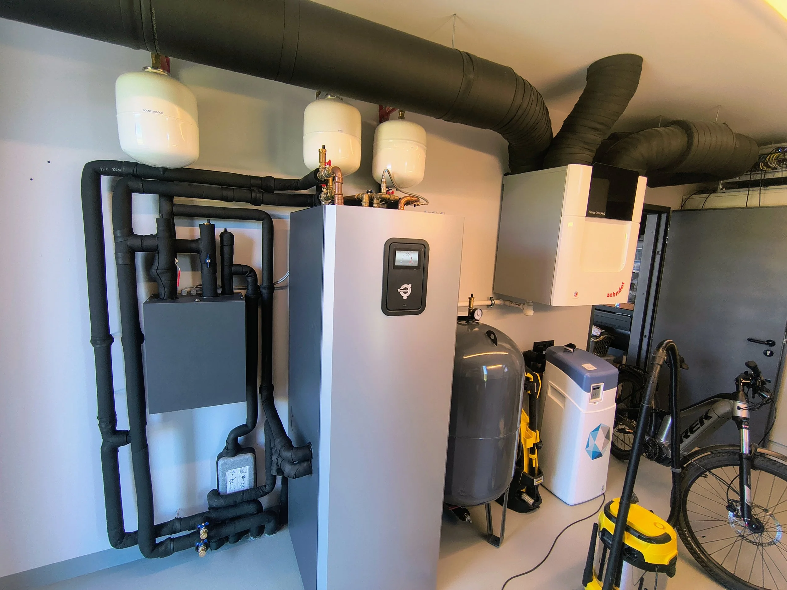 A utility room containing a boiler, pipes, and other equipment, with a bicycle and tools nearby.