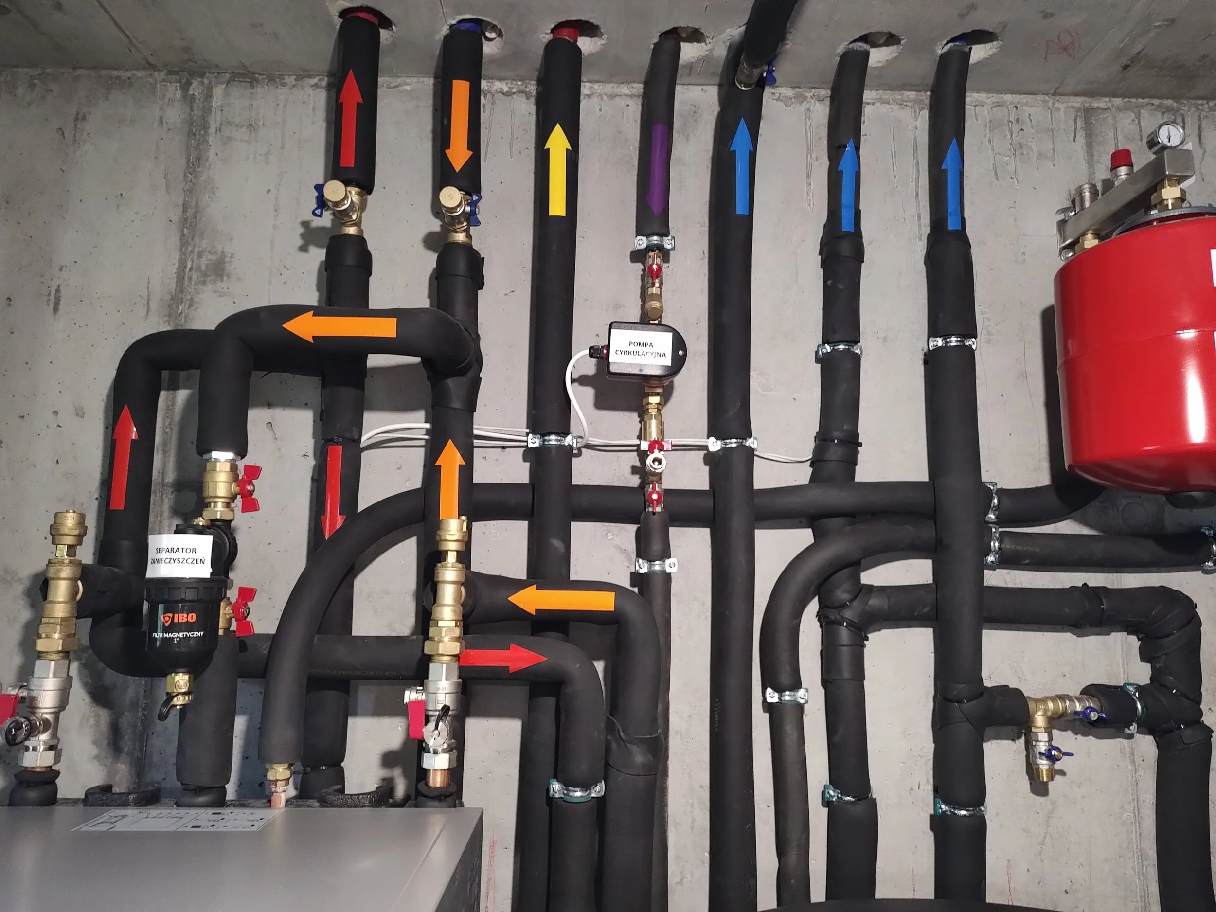 Multiple black pipes with color-coded arrows (red, orange, yellow, purple, blue) indicating flow directions, connected to a circulatory pump, mounted on a concrete wall, with various valves and fittings.