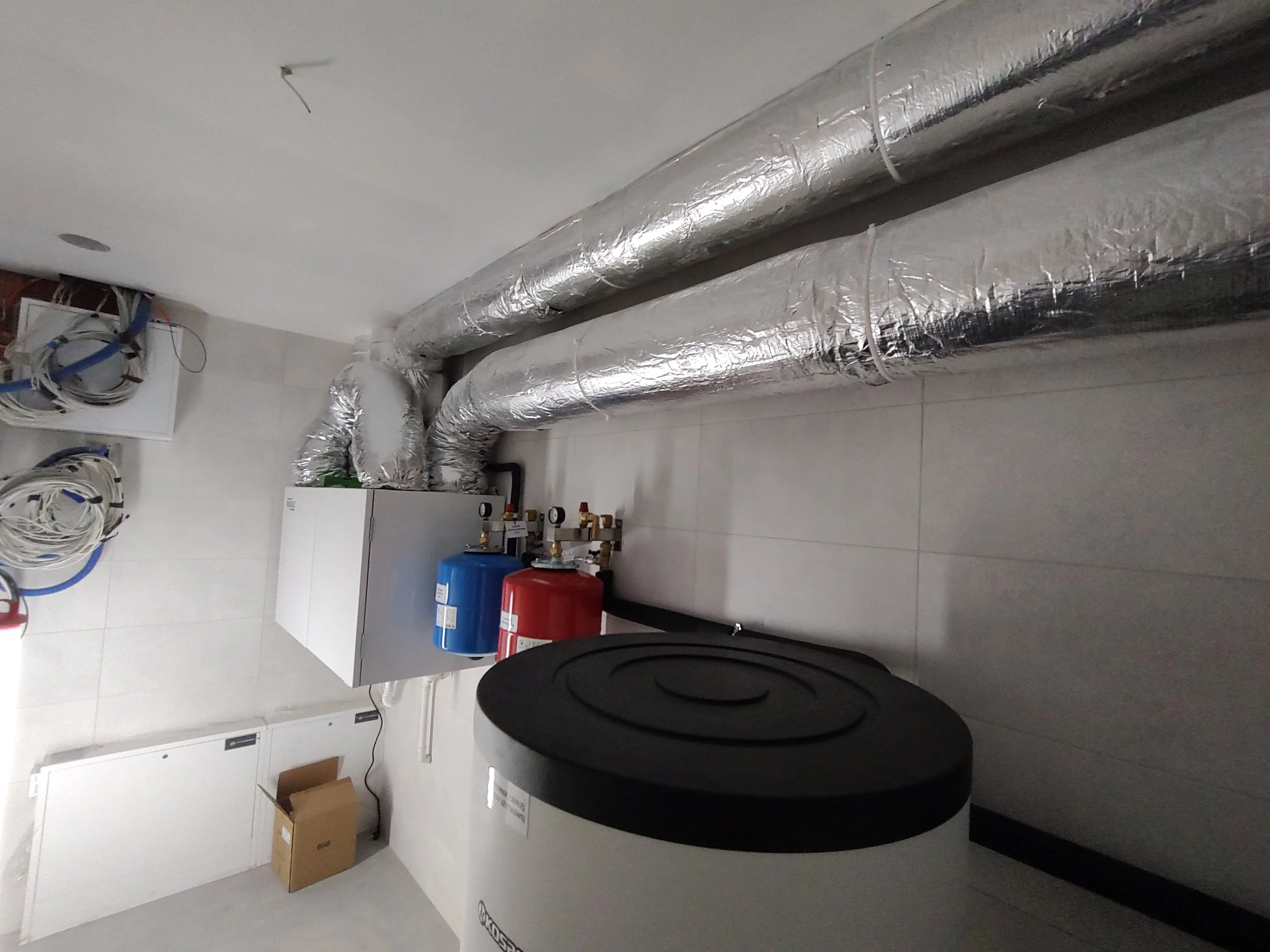 A utility or machine room showing large silver insulated ductwork, some tubing, and storage containers.