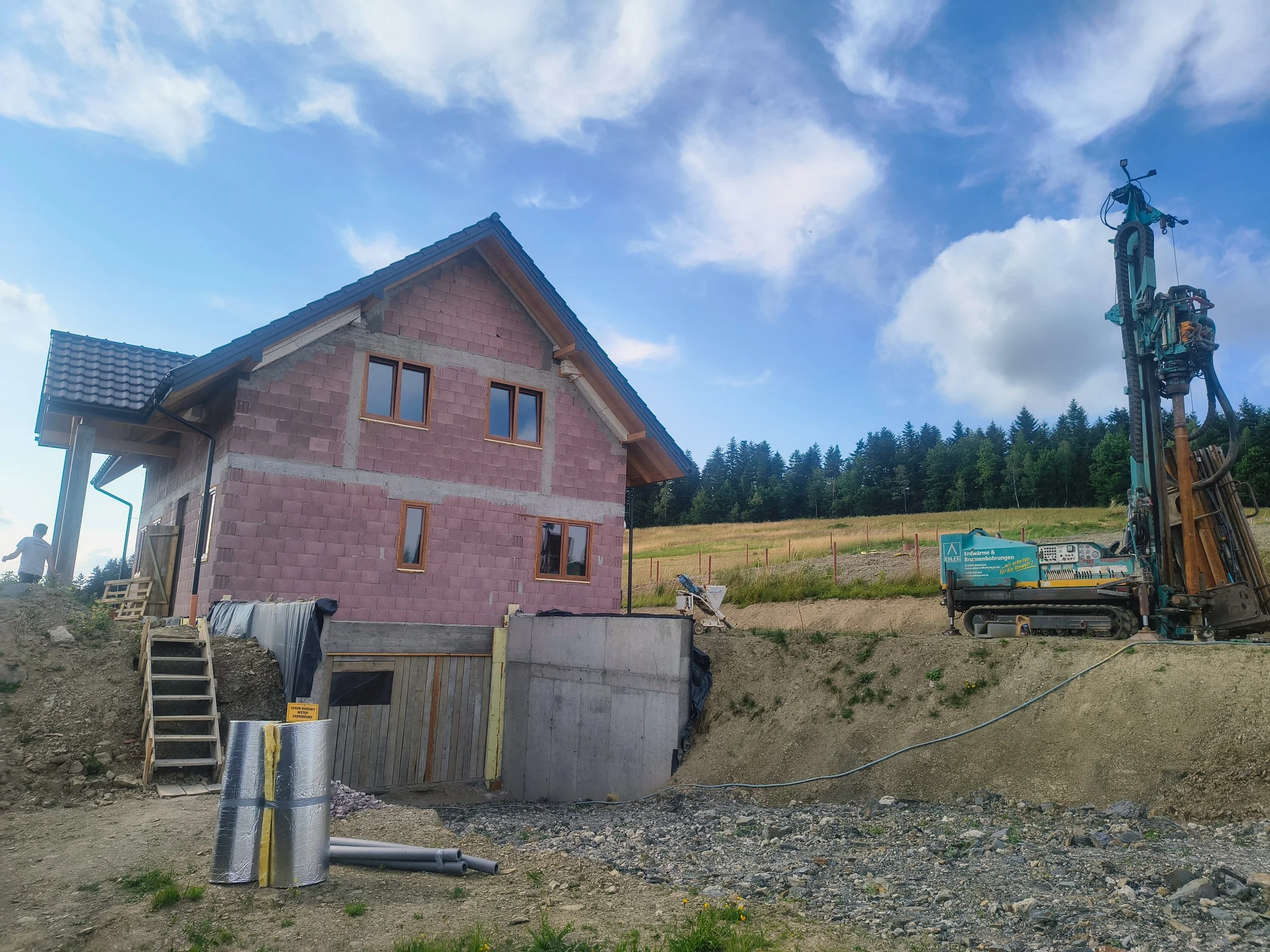 A house under construction with brick walls and a roof, next to construction machinery and pipes, on a dirt lot with a backdrop of trees and a partly cloudy sky.