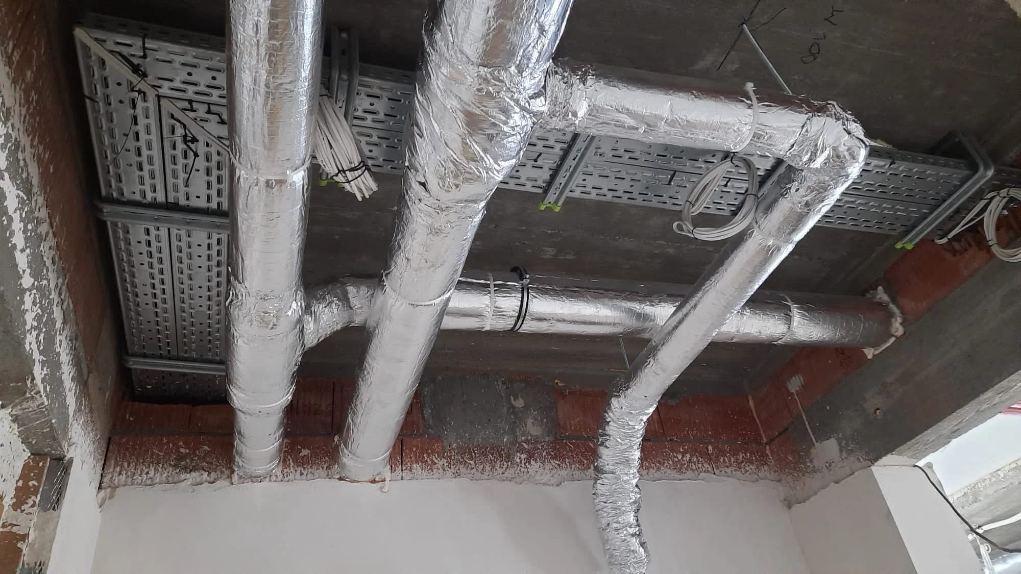 Construction site ceiling with silver insulated pipes, electrical wiring, and metal framing.