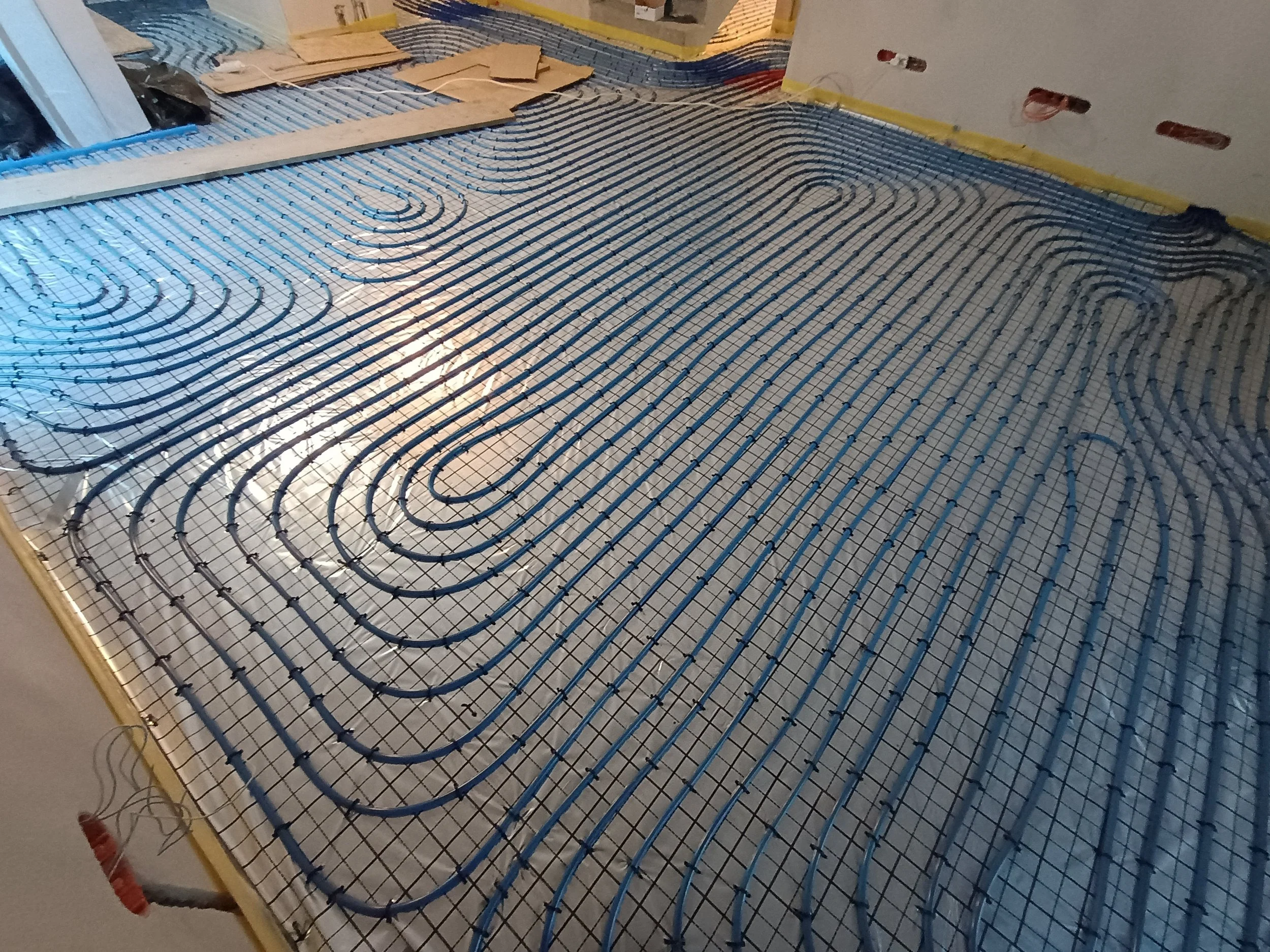 Construction site with underfloor heating pipes laid out on a wire mesh for a room. The pipes are arranged in a serpentine pattern. The room has unfinished walls and visible electrical outlets.
