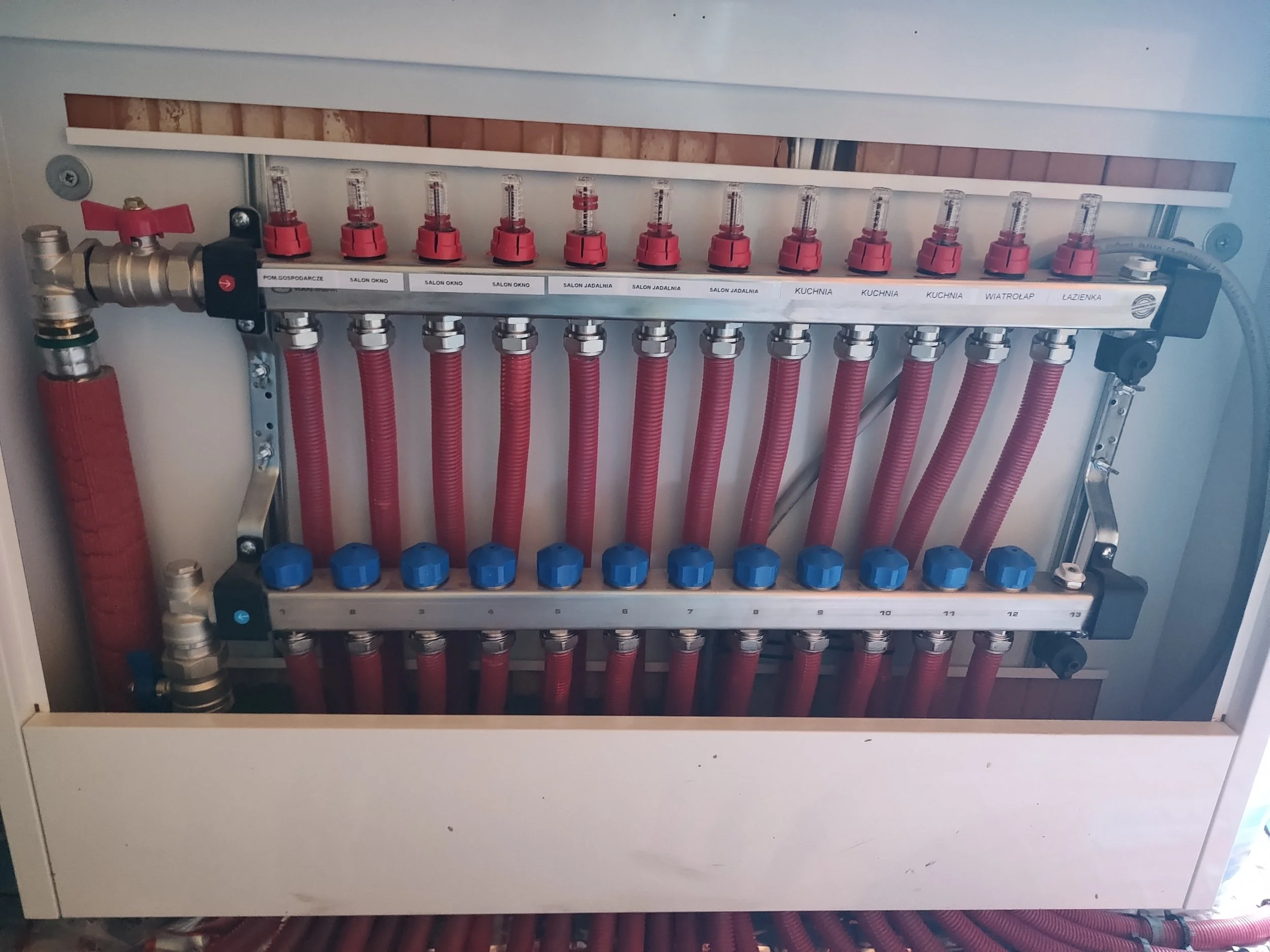 A manifold with multiple red pipes connected to it, labeled with room names in Polish, such as "Salon Okno" and "Kuchnia".