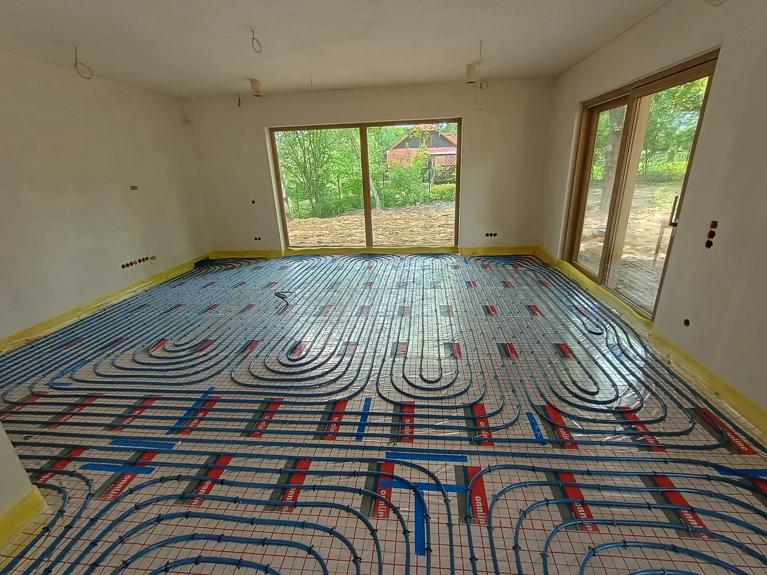 Inside a room under construction showing radiant floor heating pipes installed over a mesh on the floor, with large windows revealing a green outdoor scene and a house in the background.