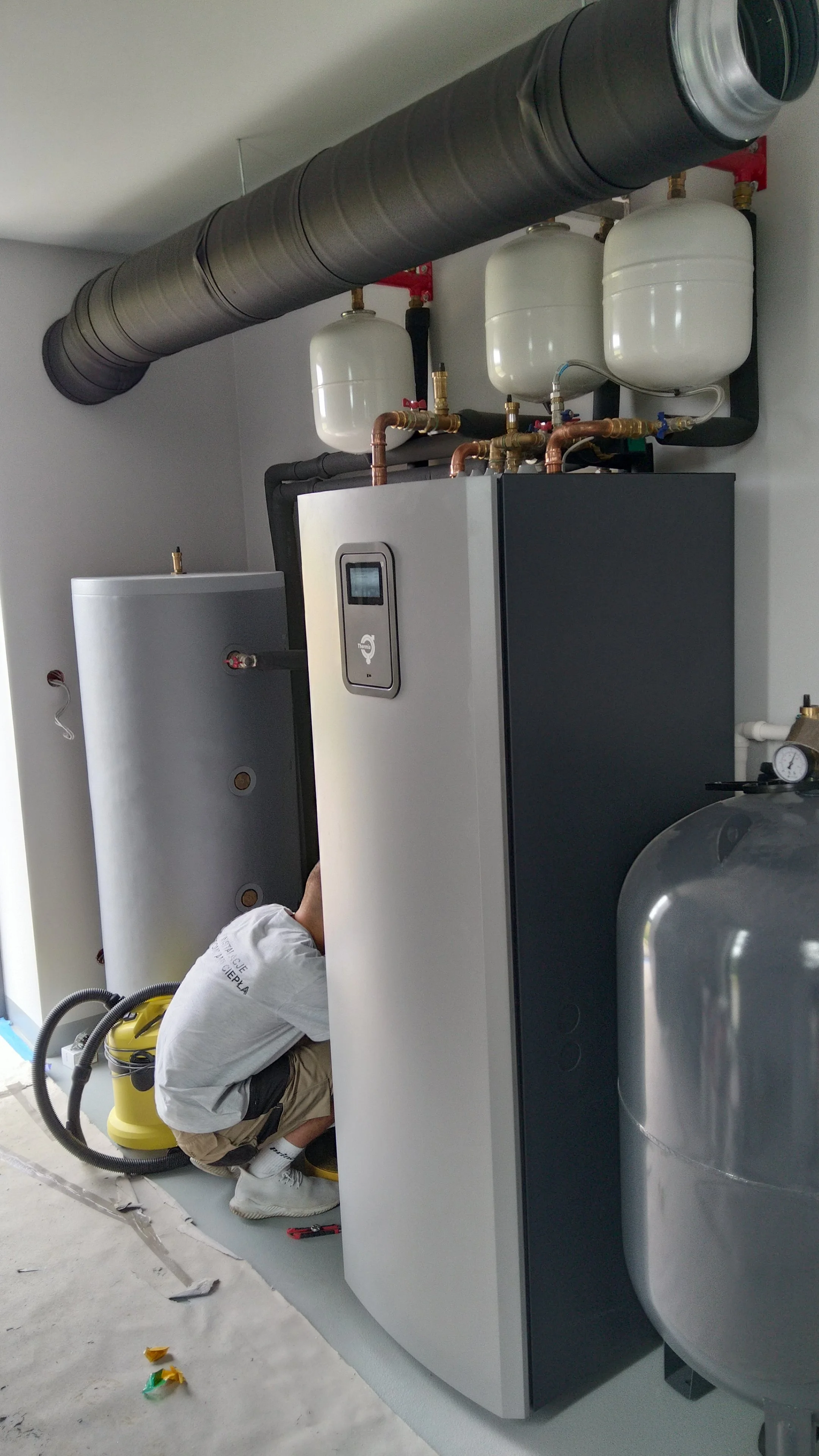 Technician installing or maintaining a large industrial or commercial HVAC system with two white expansion tanks, copper pipes, and a black ventilation duct.