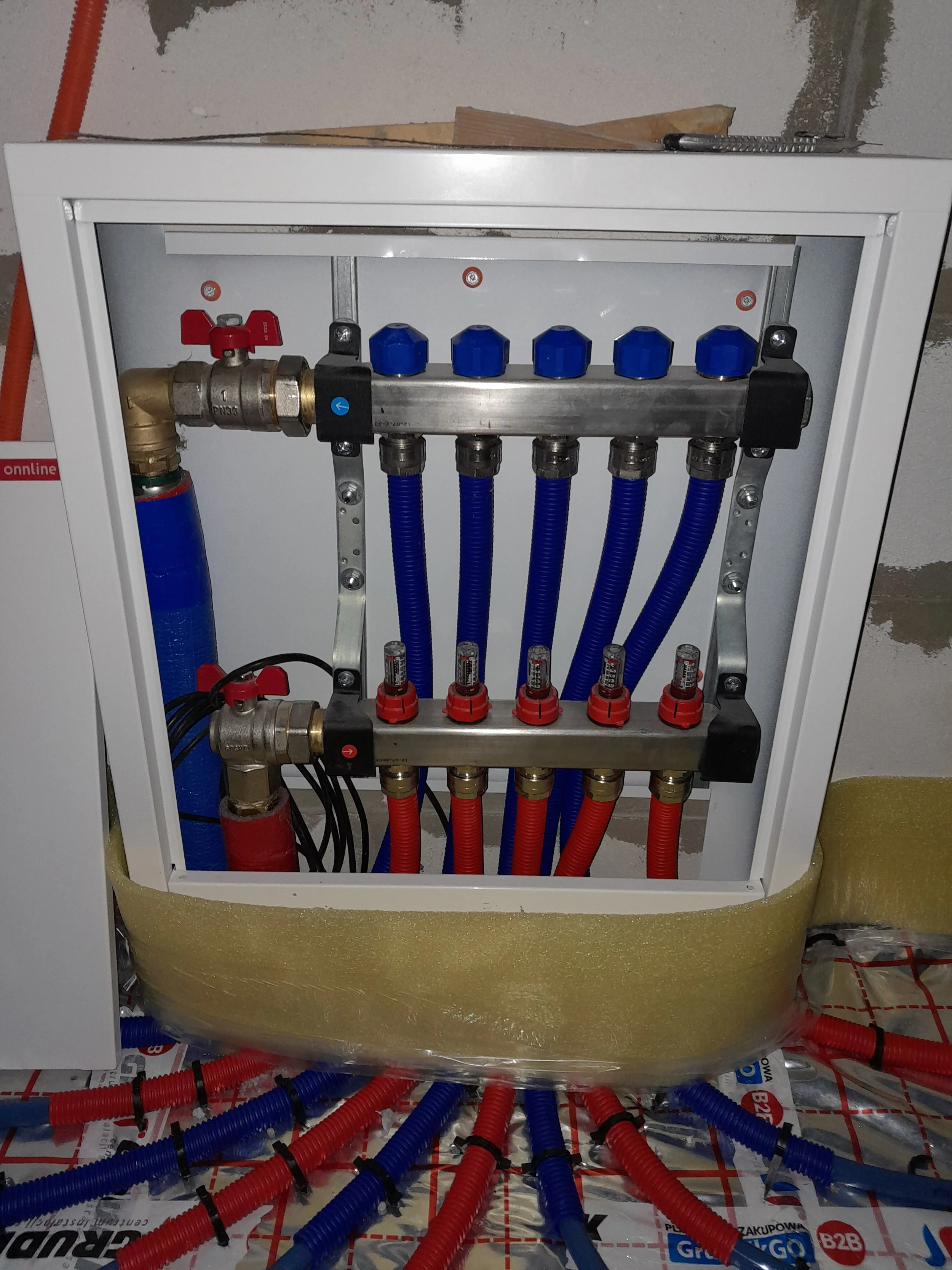 A plumbing manifold with several blue and red pipes connected, installed inside a white electrical box on a wall. The pipes are organized with metal brackets and the setup appears to be part of a plumbing system under construction or installation.