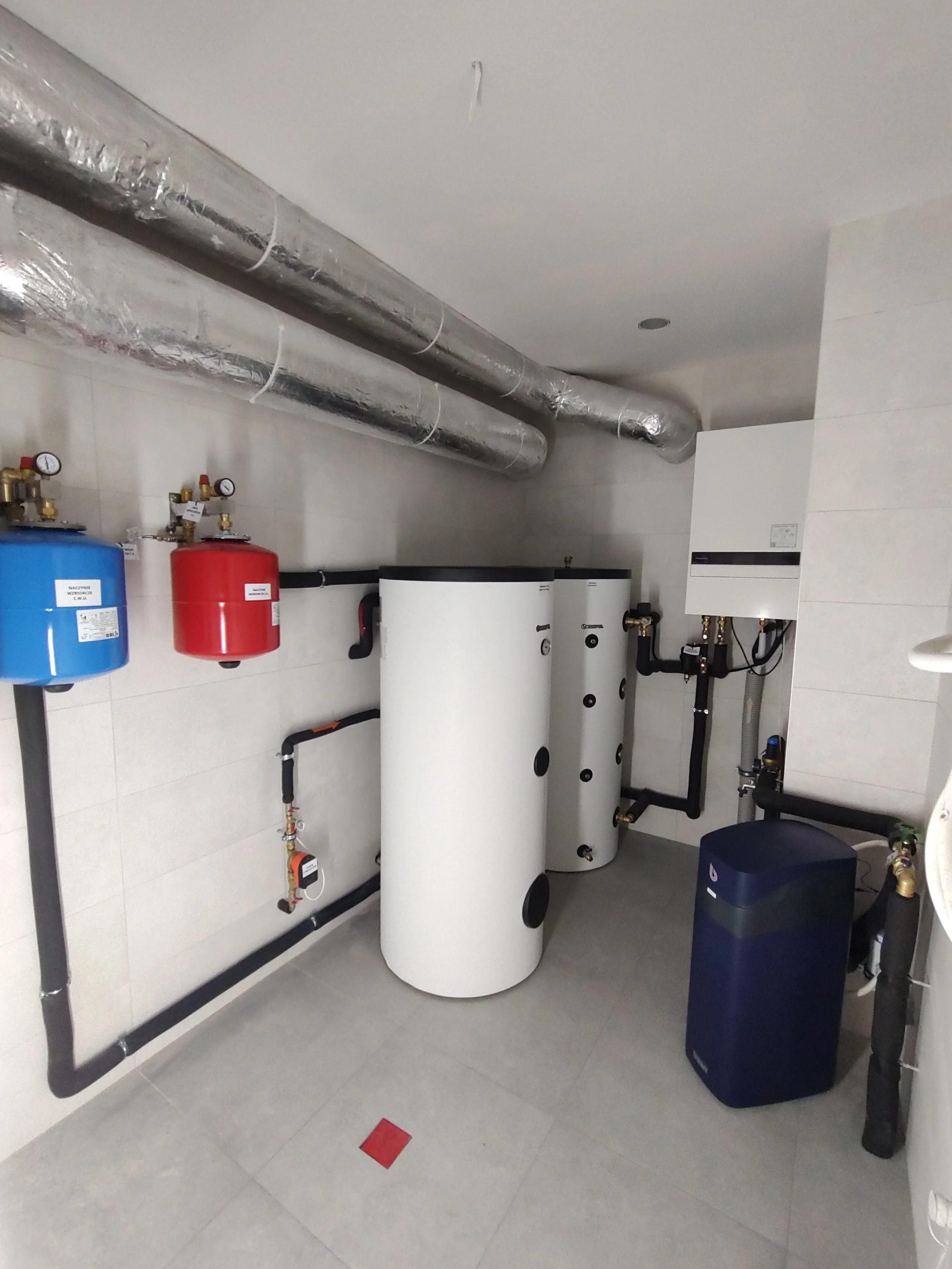 Room with two large white water heater tanks, insulated silver pipes running along the ceiling and wall, black pipes, a blue filter, and a red and black pressure tank inside a building with tiled walls and floor.