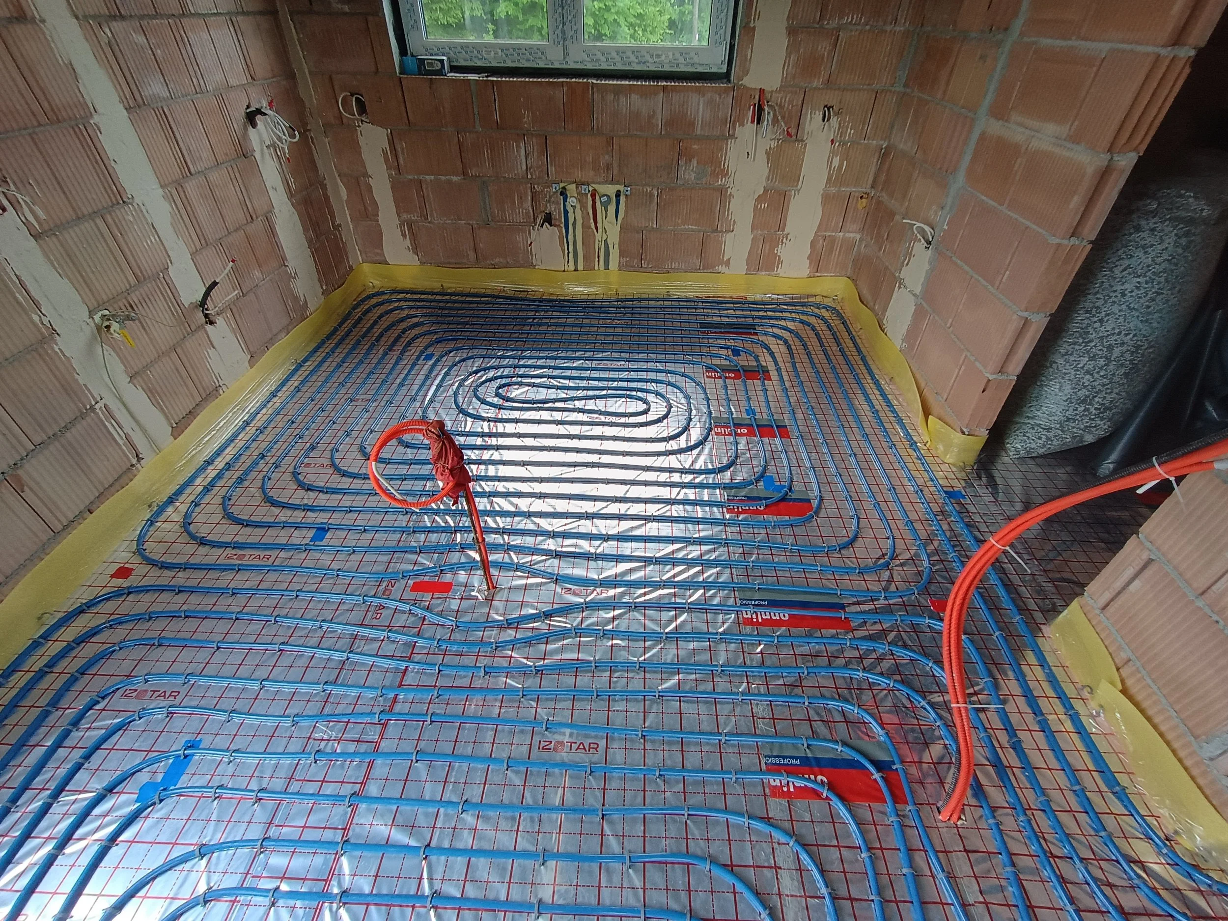 Interior of a room under construction with an underfloor heating system installed. Blue pipes are laid out in a serpentine pattern on a reflective insulation layer, with exposed brick walls and a small window showing greenery outside.