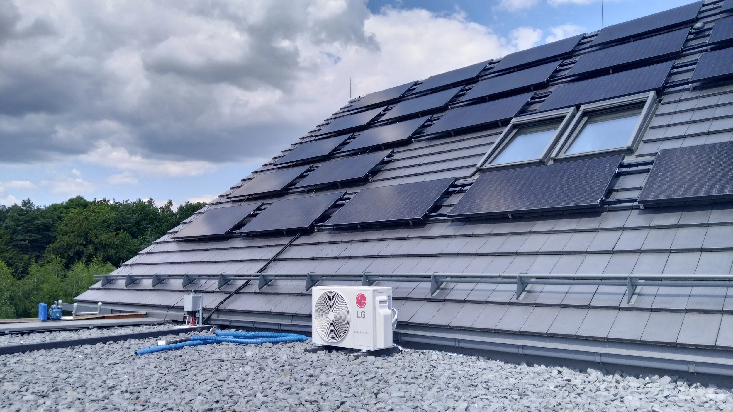 Rooftop with multiple solar panels installed, air conditioning unit, and gravel surface, with trees and cloudy sky in the background.
