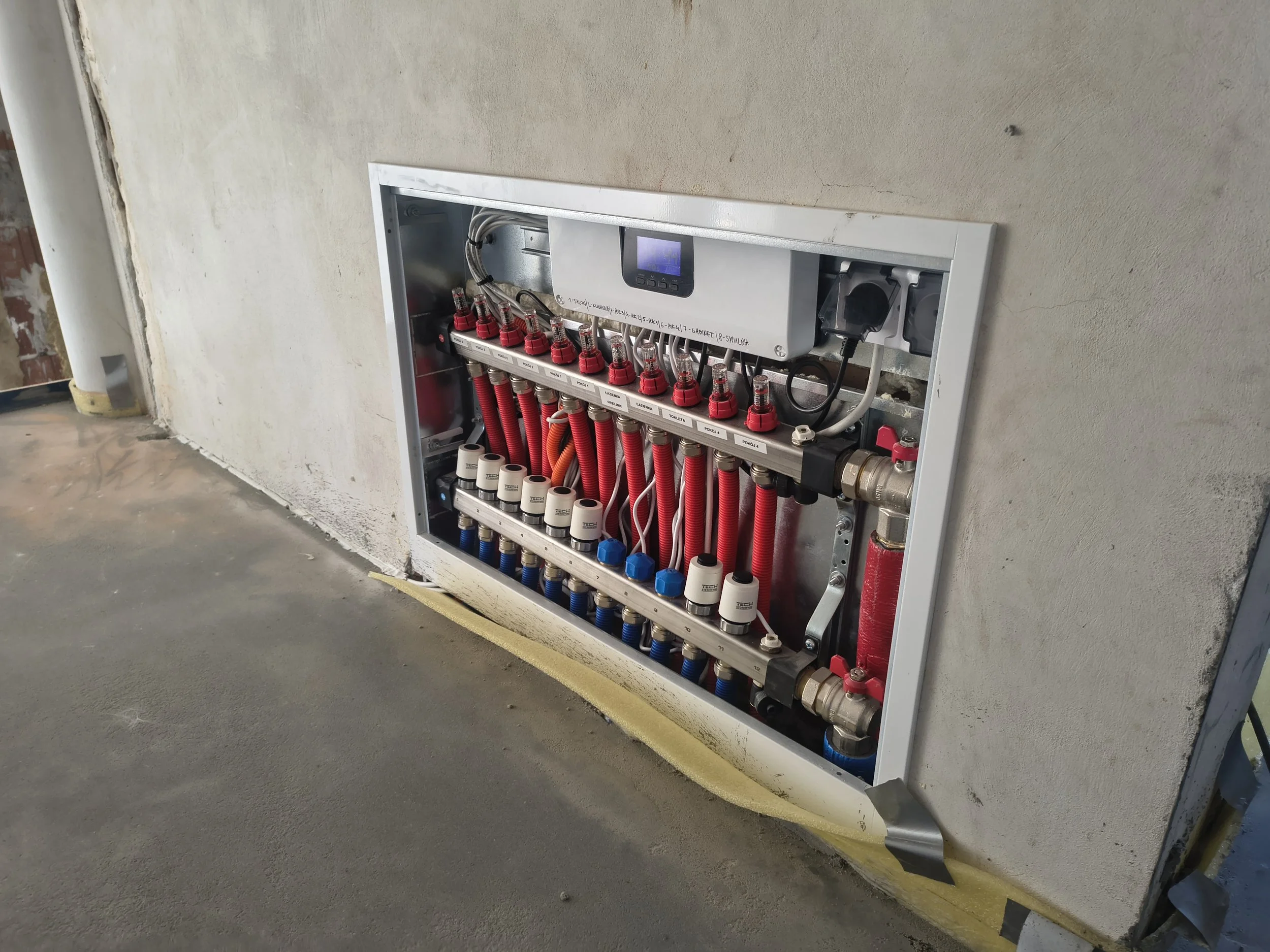 Open electrical panel with organized red, blue, and white pipes, a small digital display, and various wiring, installed on a concrete wall in a construction site.