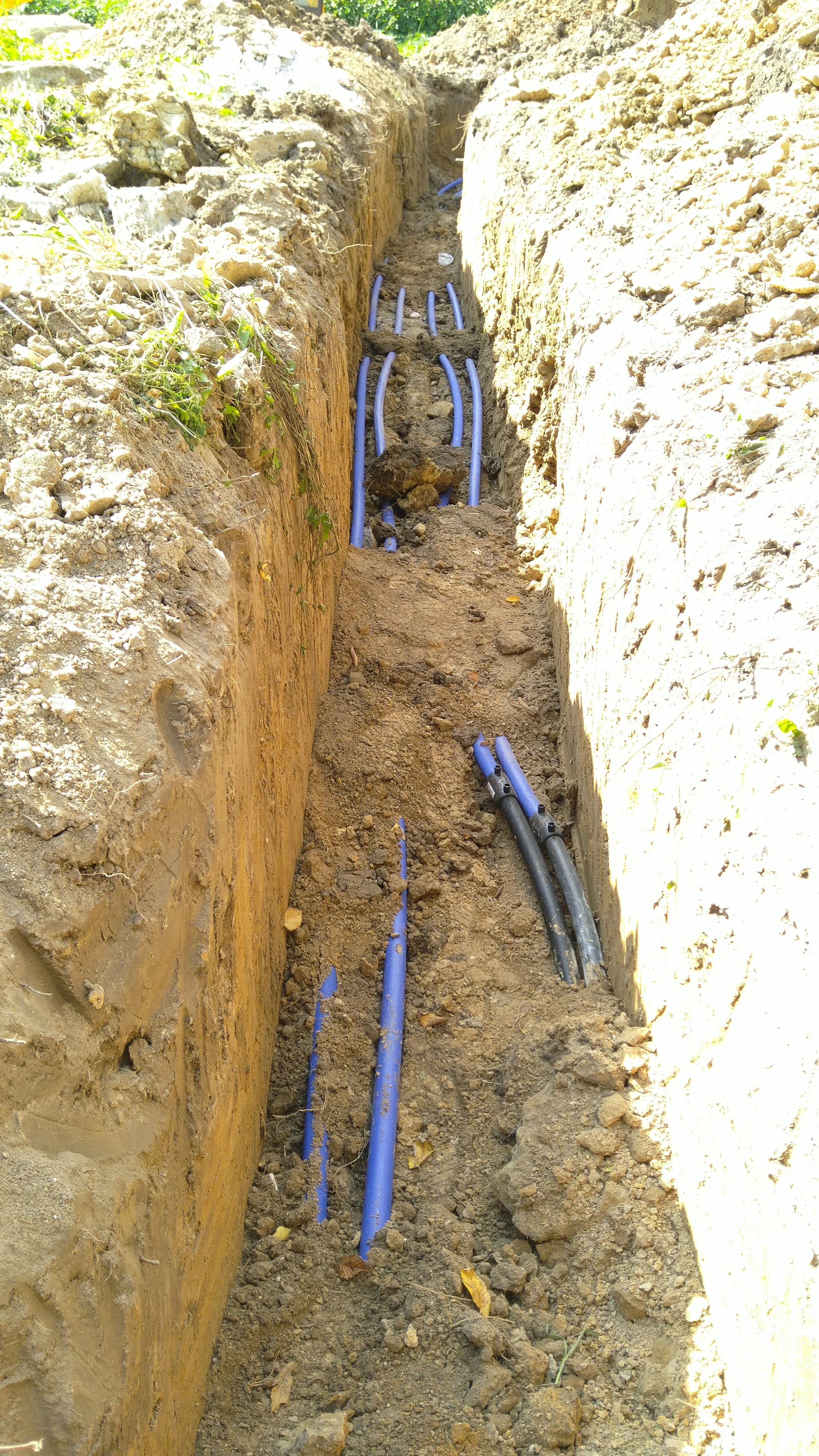 Underground utility pipes, some blue and one black, are being installed in a narrow trench with soil on both sides.