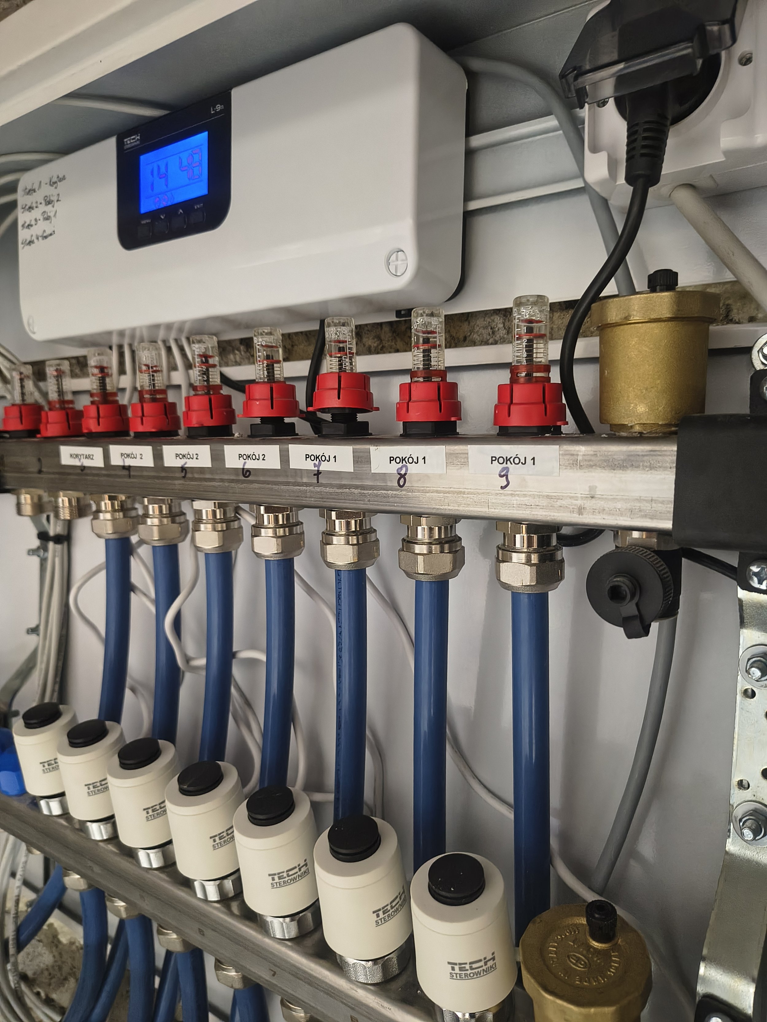 Close-up of a mechanical control panel with blue digital display showing 14.9. There are multiple numbered valves with red and clear caps, connected to blue pipes. Labels underneath include 'KORYTARZ', 'POKÓJ 2', and 'POKÓJ 1'. Additional components 