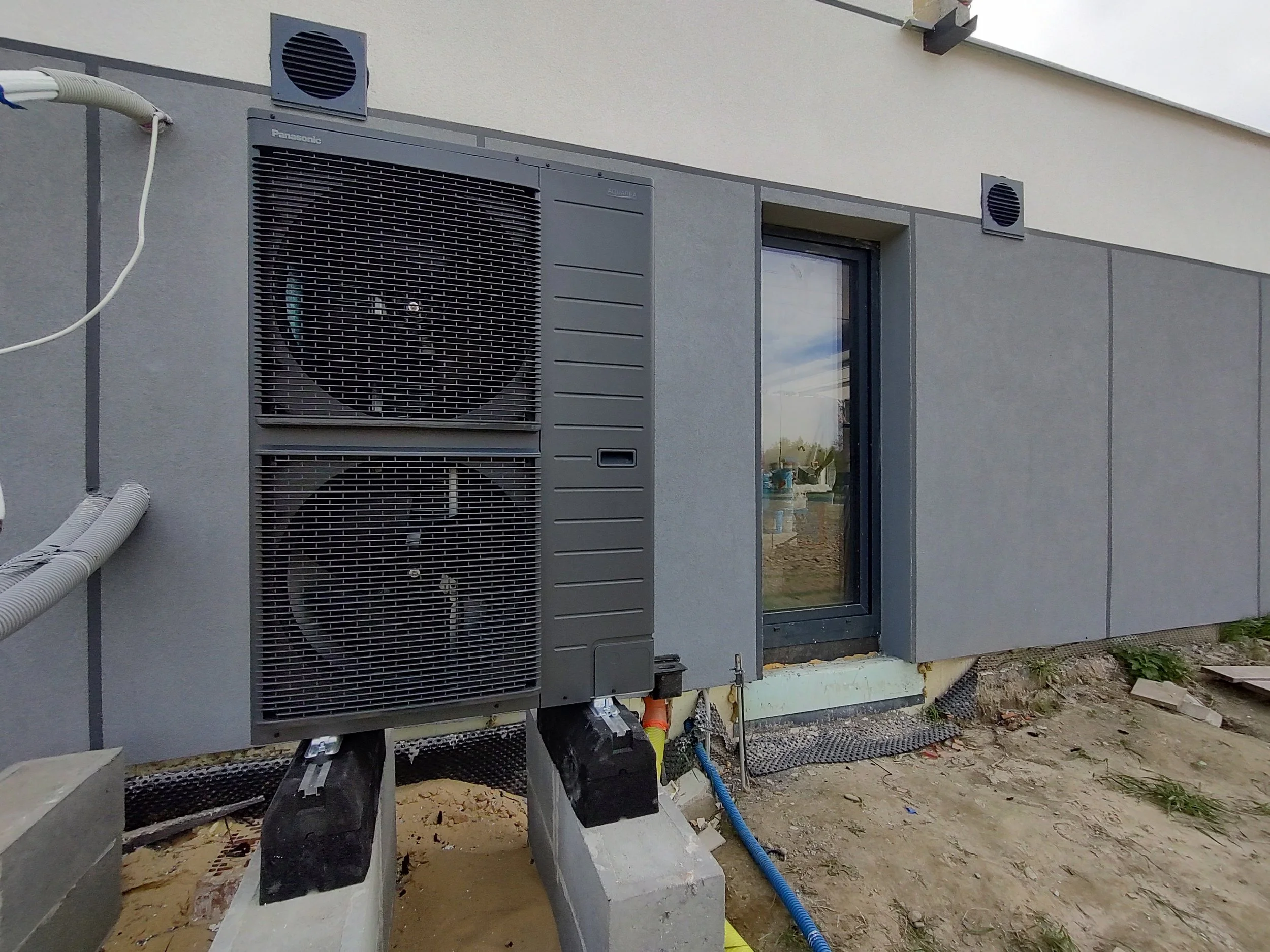 Exterior of a building with an HVAC system installed on the wall. The system includes two large fans and vents, with cables and pipes connected to it, and a window nearby reflecting the outdoors.
