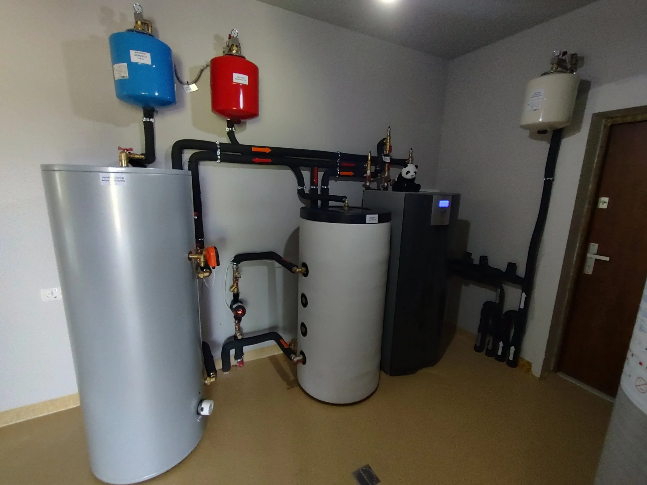 A utility room with a large cylindrical water heater, pipes, and pressure tanks on the wall, along with a small control panel.