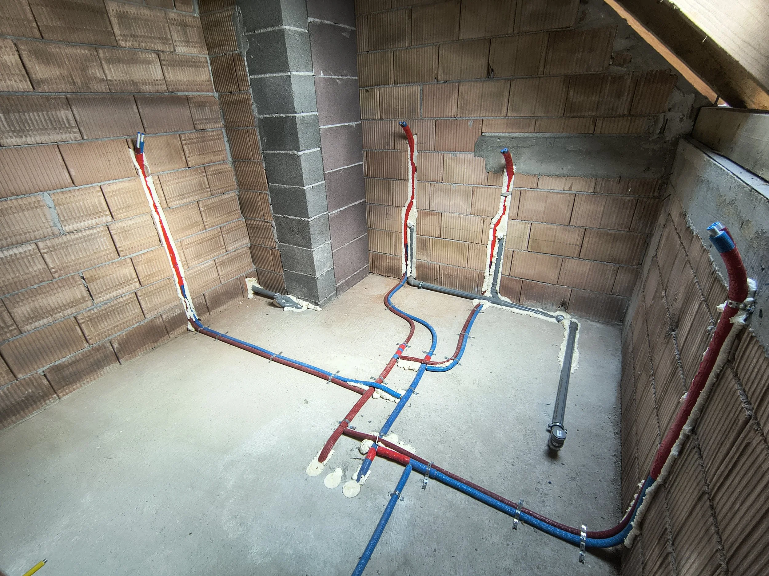 This is an indoor construction site with exposed brick walls and plumbing pipes. The pipes are red and blue, indicating hot and cold water lines, and are installed along the floor and up the wall. Some pipes are wrapped with insulation material. The 