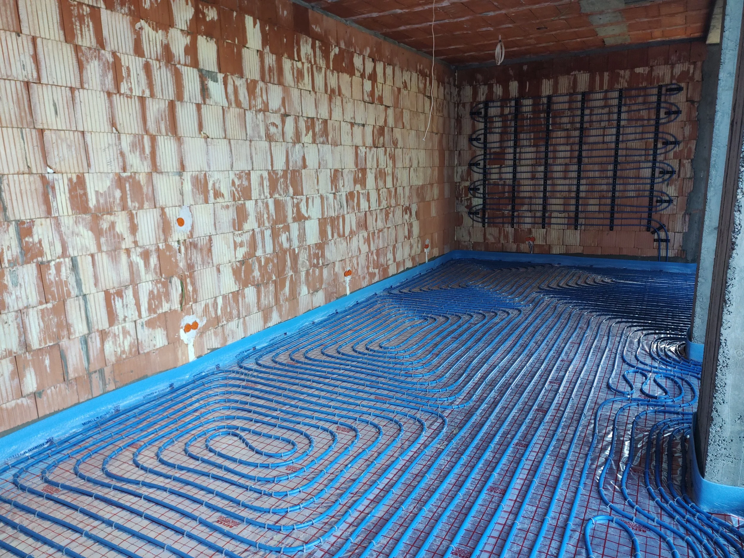Interior of a building under construction, showing brick walls and a floor with underfloor heating pipes installed and ready for covering.