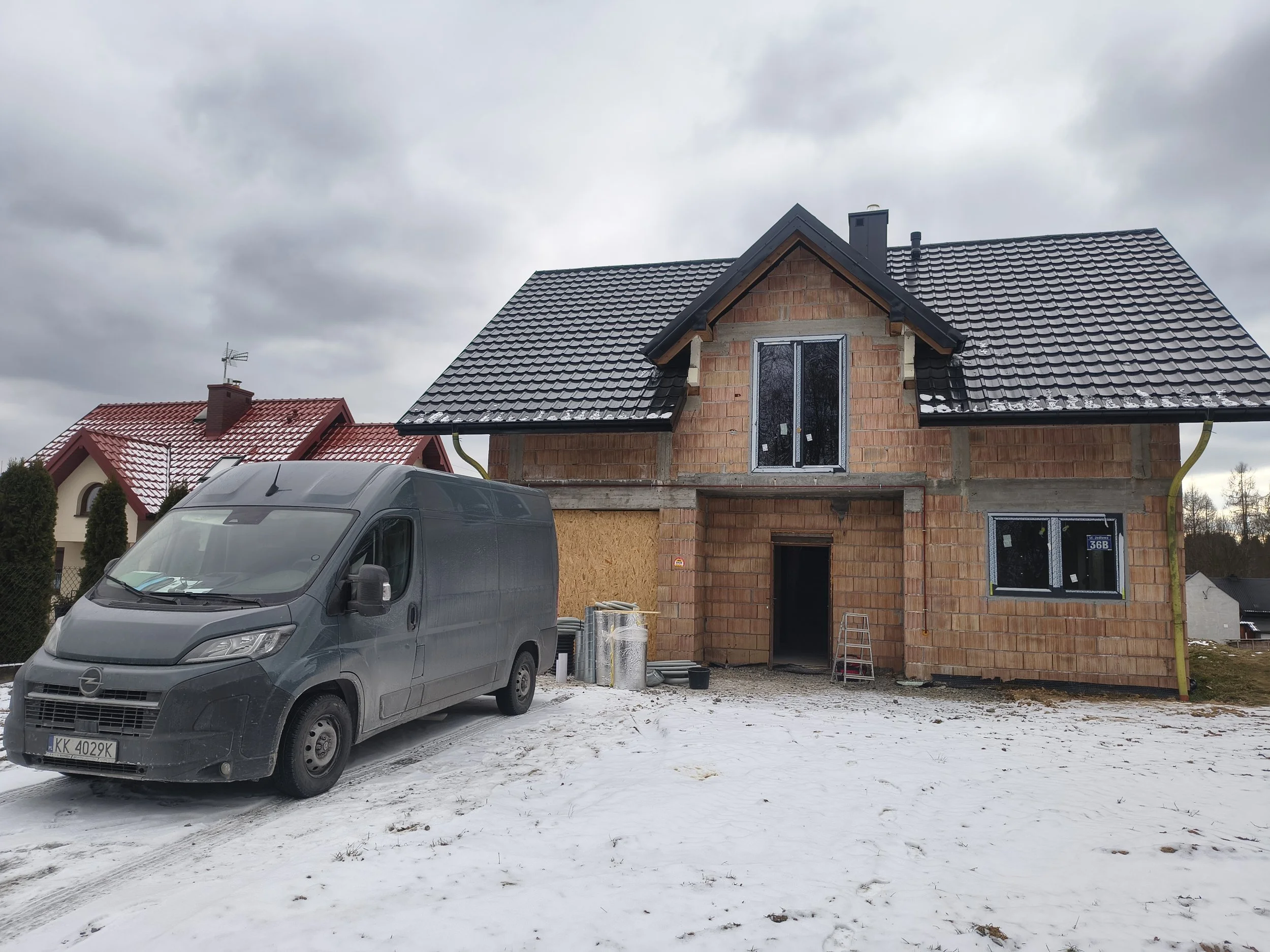 A house under construction with brick walls and a black tile roof, surrounded by a snowy yard, with a gray van parked in front.
